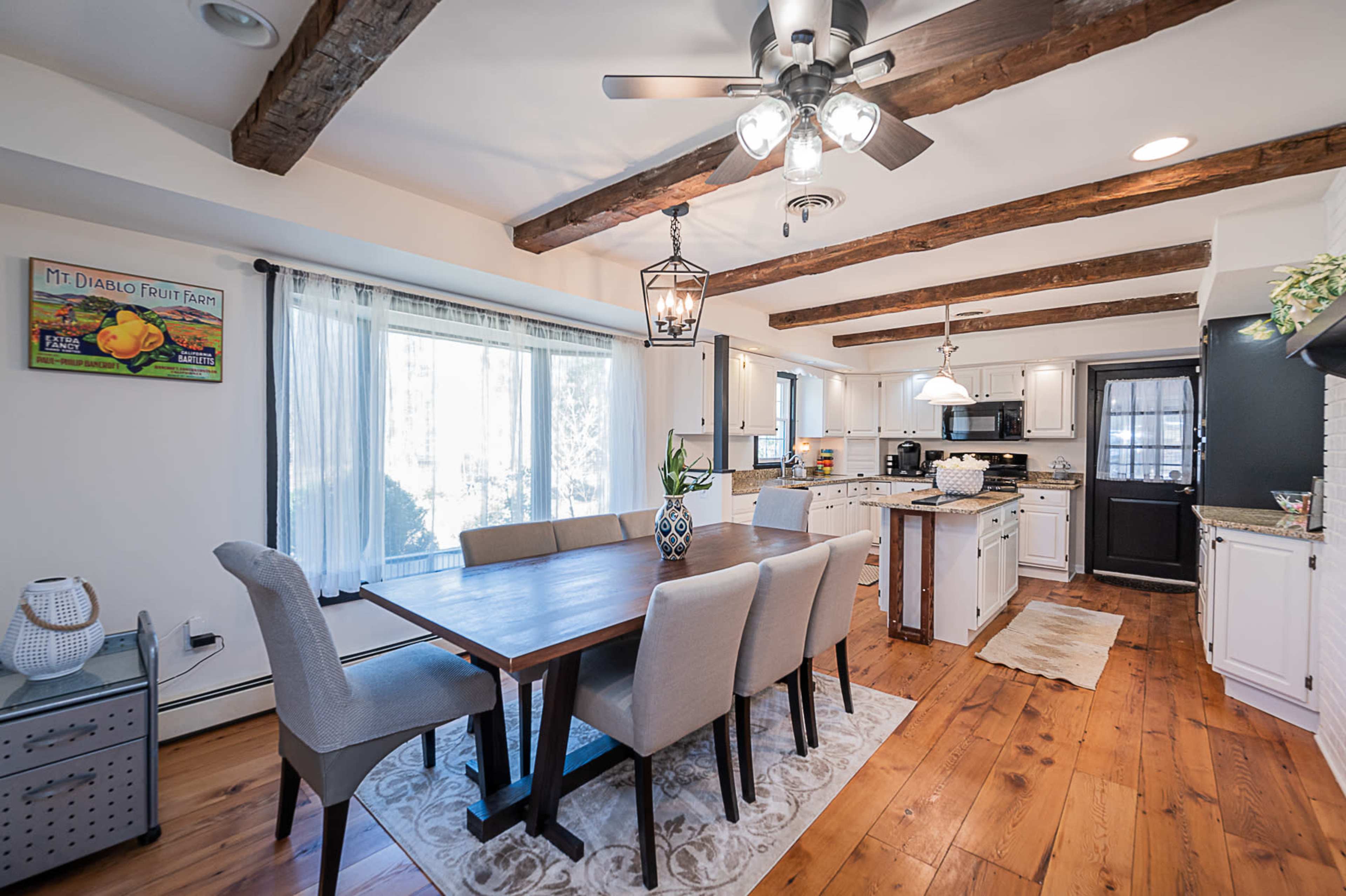A bright kitchen and dining area with wooden beams on the ceiling, a long dining table surrounded by upholstered chairs, and modern kitchen appliances.