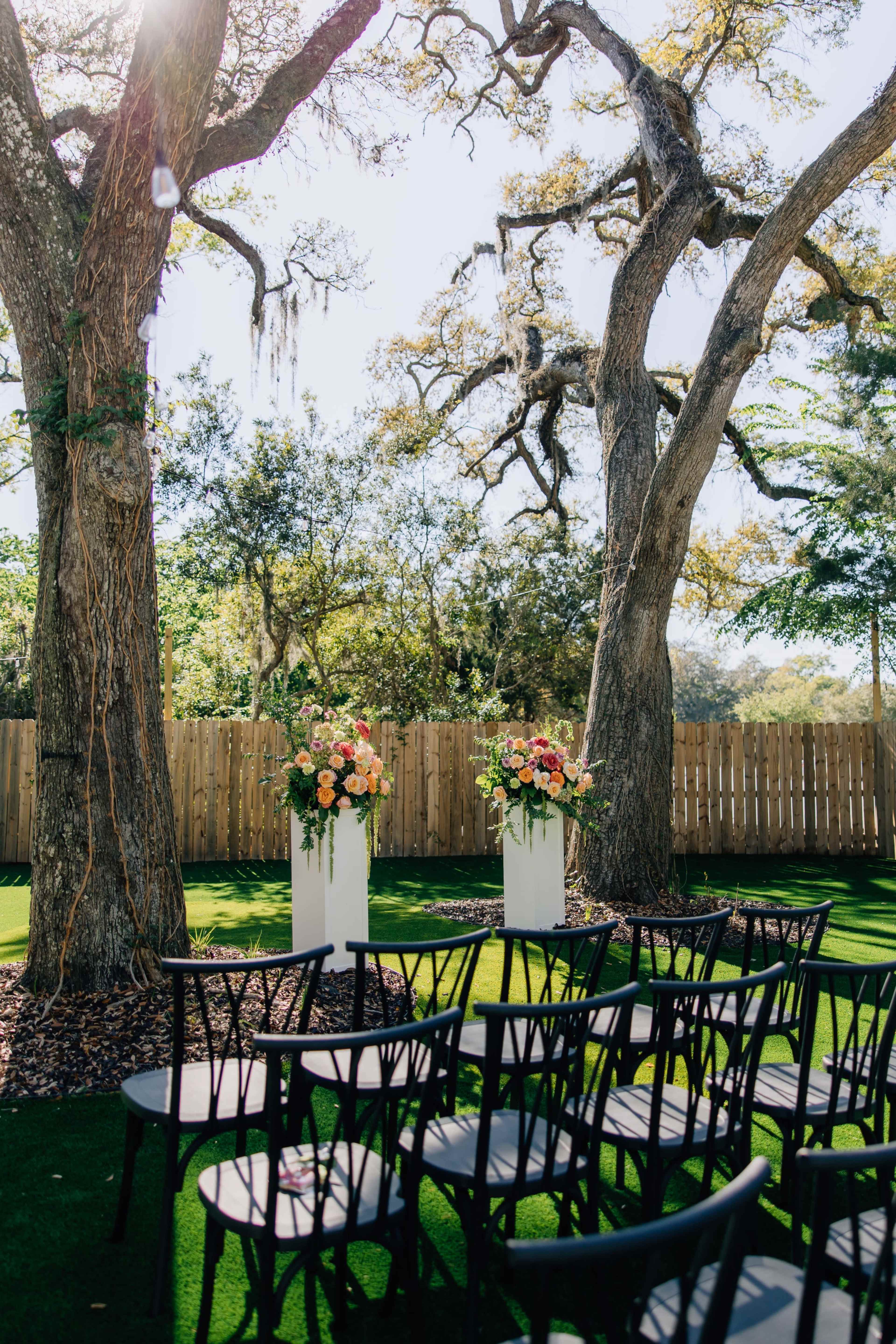 The image shows a garden setting with two large floral arrangements on white pedestals among wooden chairs placed on green grass.