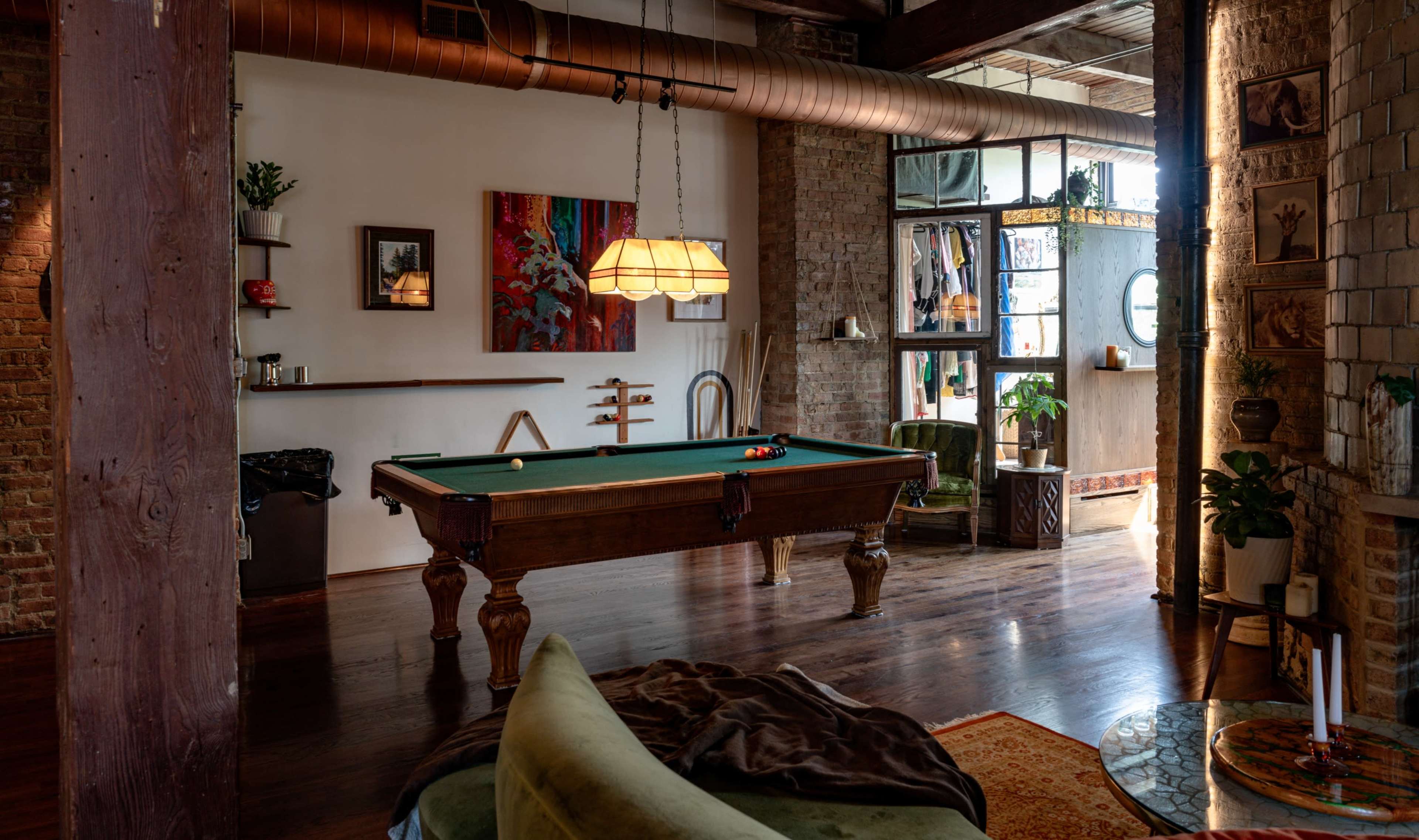 The image shows a cozy interior space featuring a billiards table, wooden beams, a green sofa, and large windows with natural light.