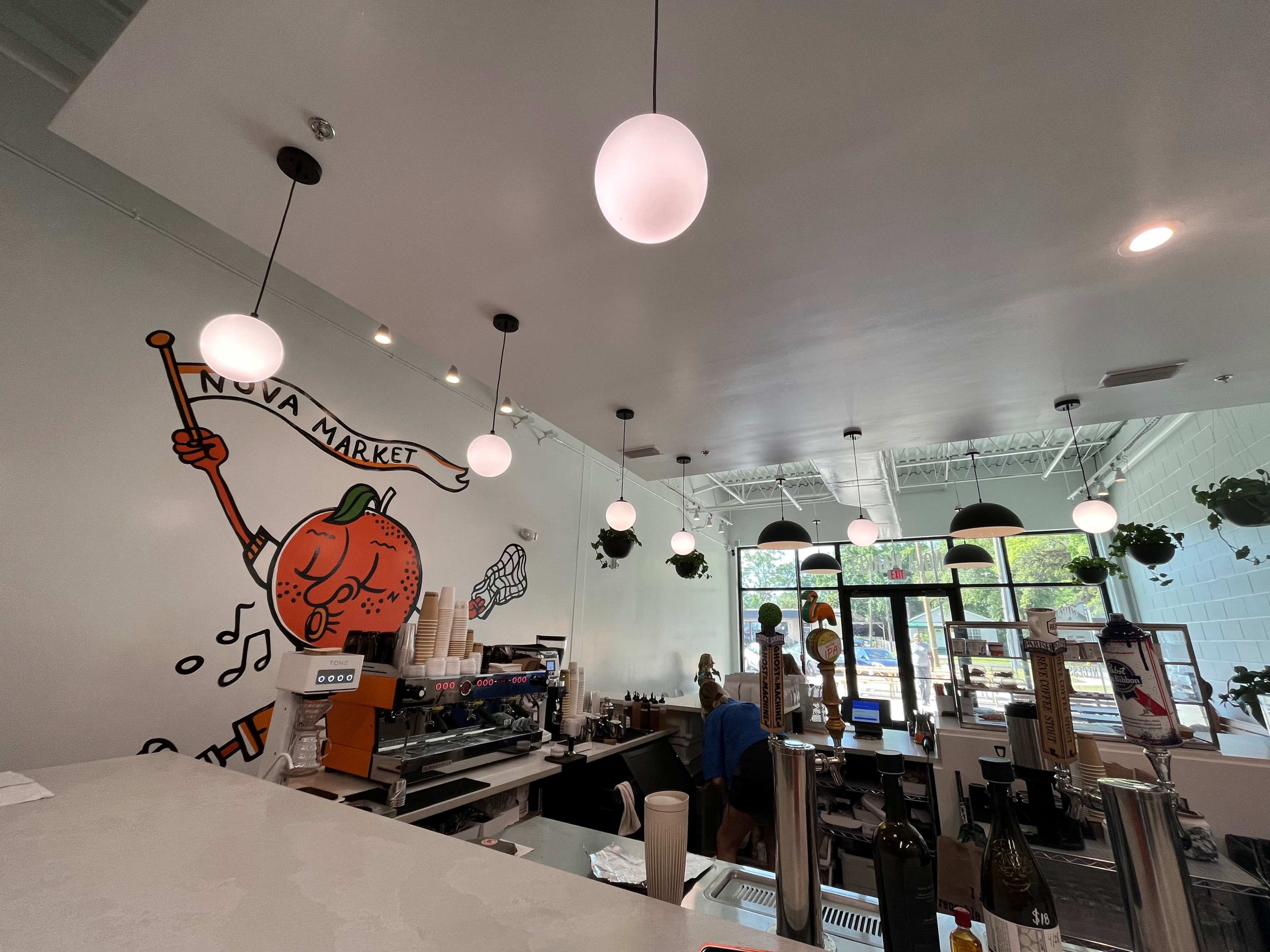 The interior of a café features a counter with a coffee machine, hanging lights, and a mural of a character holding a musical instrument.
