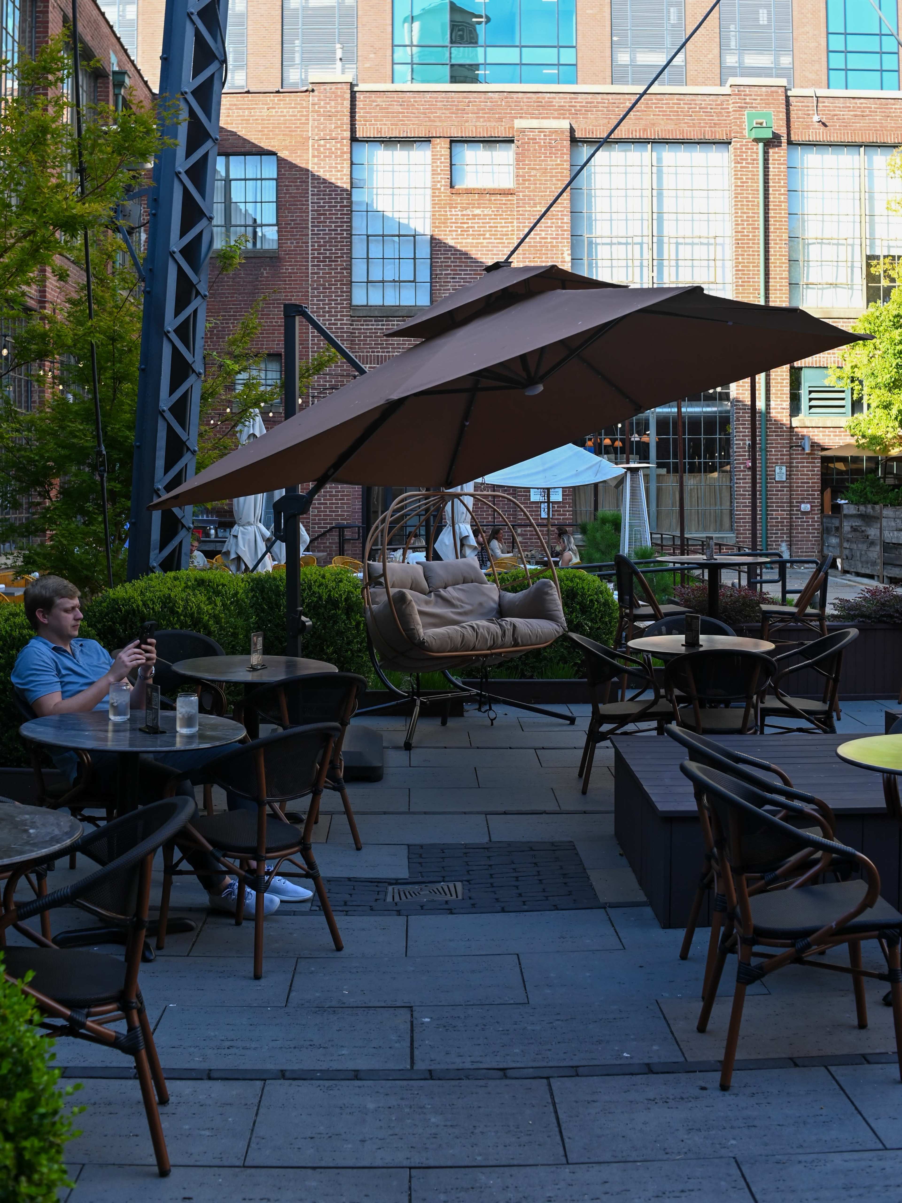 A young man sits at a patio table, using his phone while a large umbrella and a hanging chair are visible in the background.