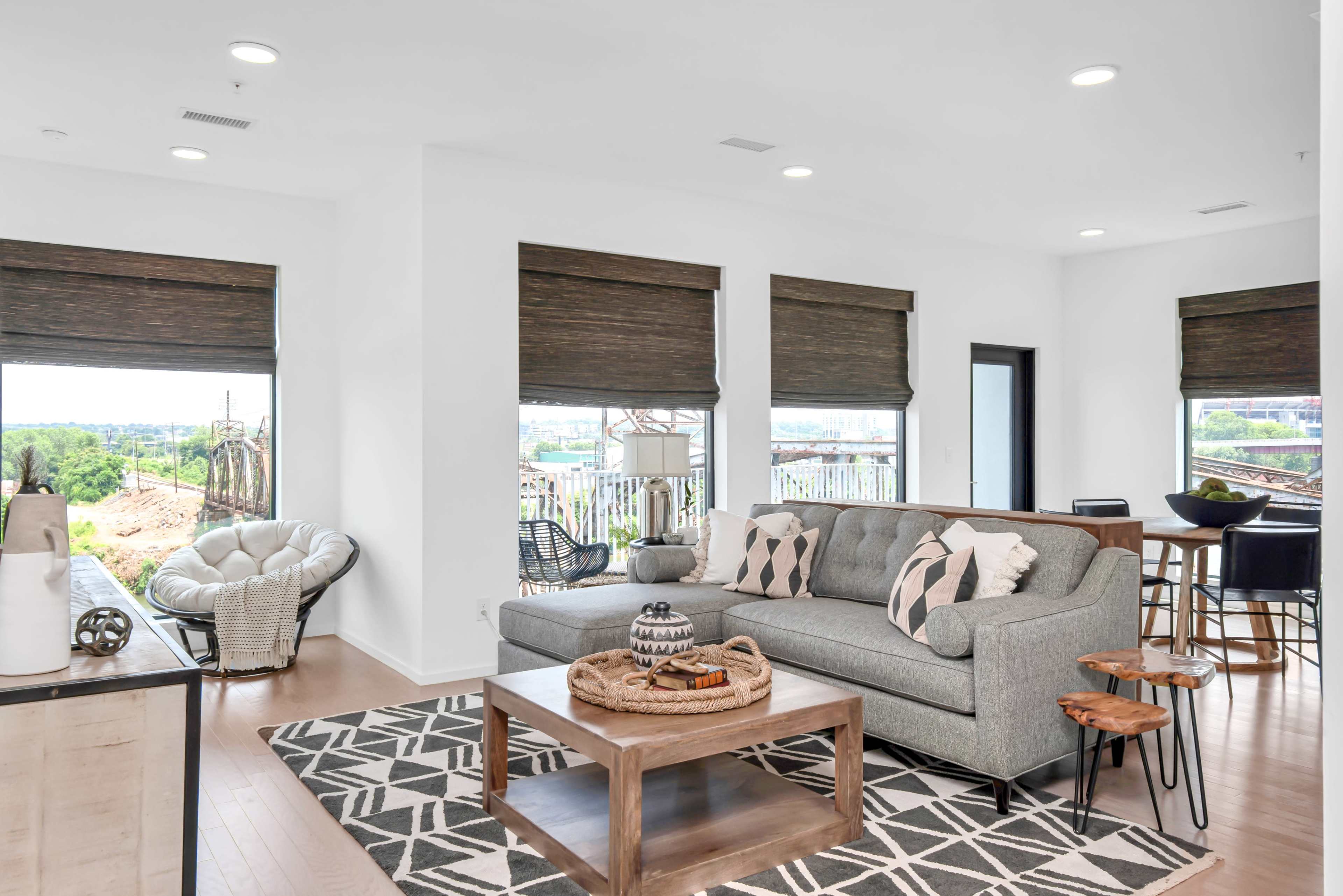 A modern living room features a gray sofa, wooden coffee table, and patterned rug, with large windows showing a view of the outdoors.