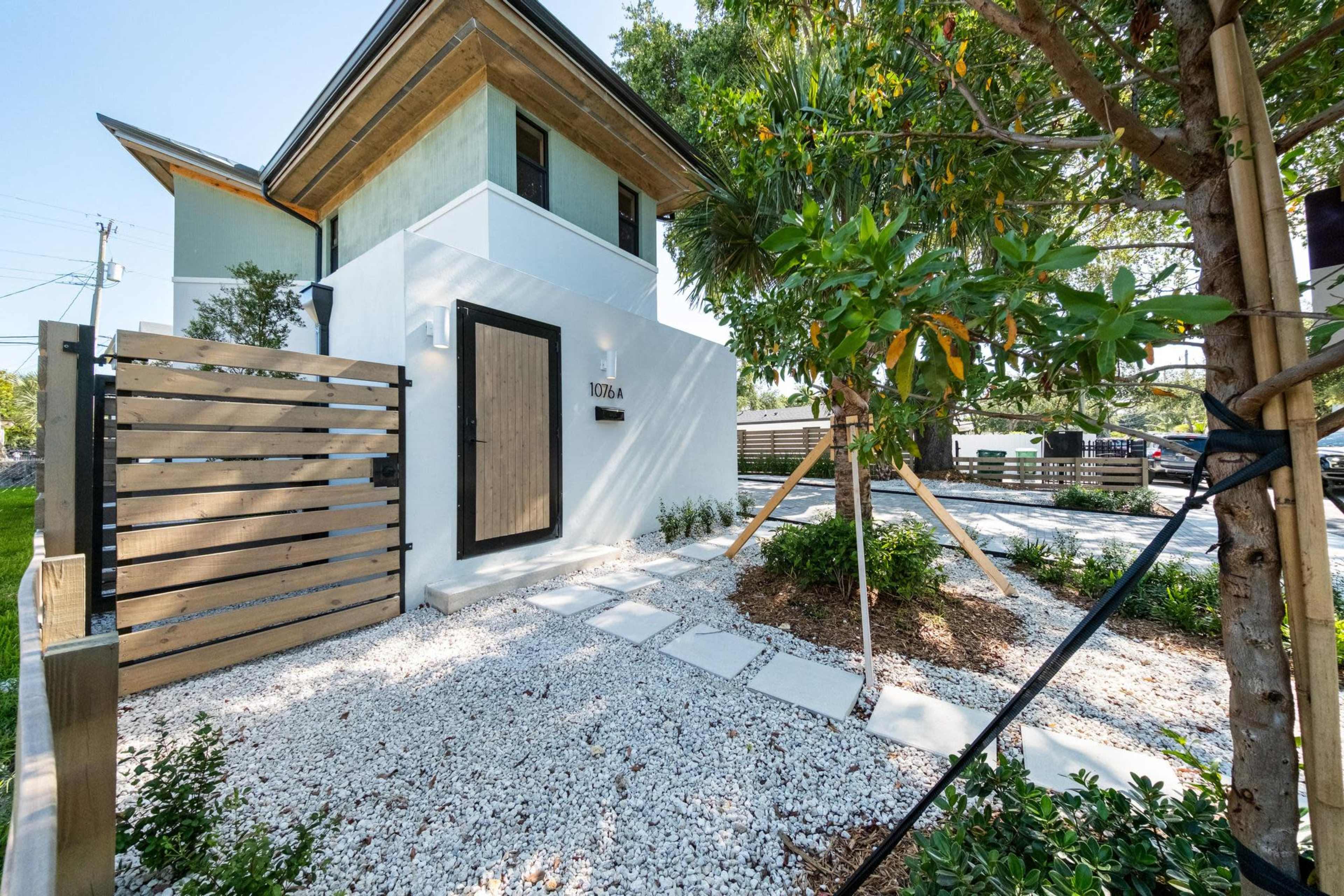 The image shows a modern house with a wooden door, surrounded by a landscaped garden featuring white gravel pathways and lush greenery.
