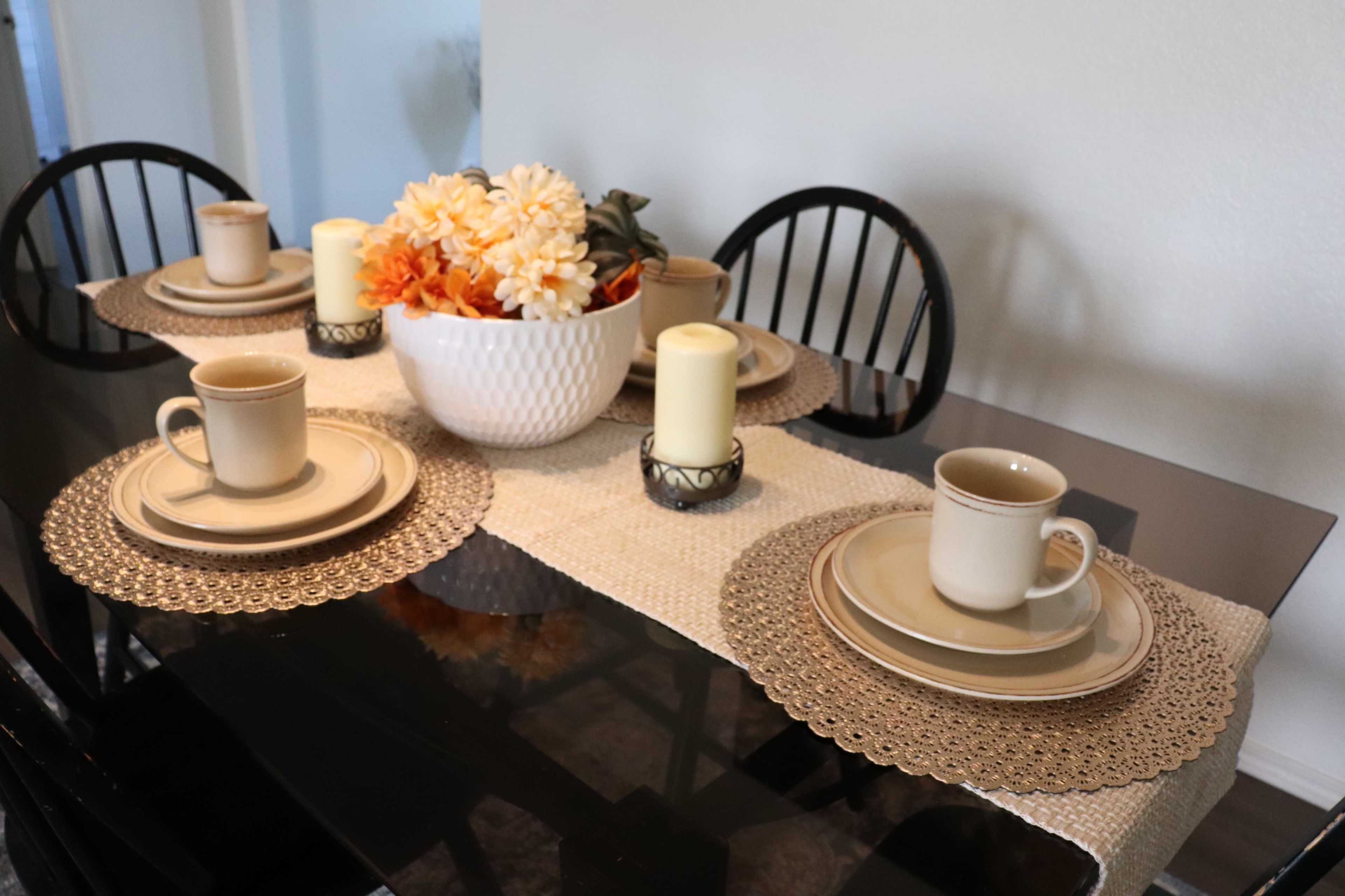 A dining table is set with beige plates and mugs, adorned with a floral centerpiece and lit candles on a runner.