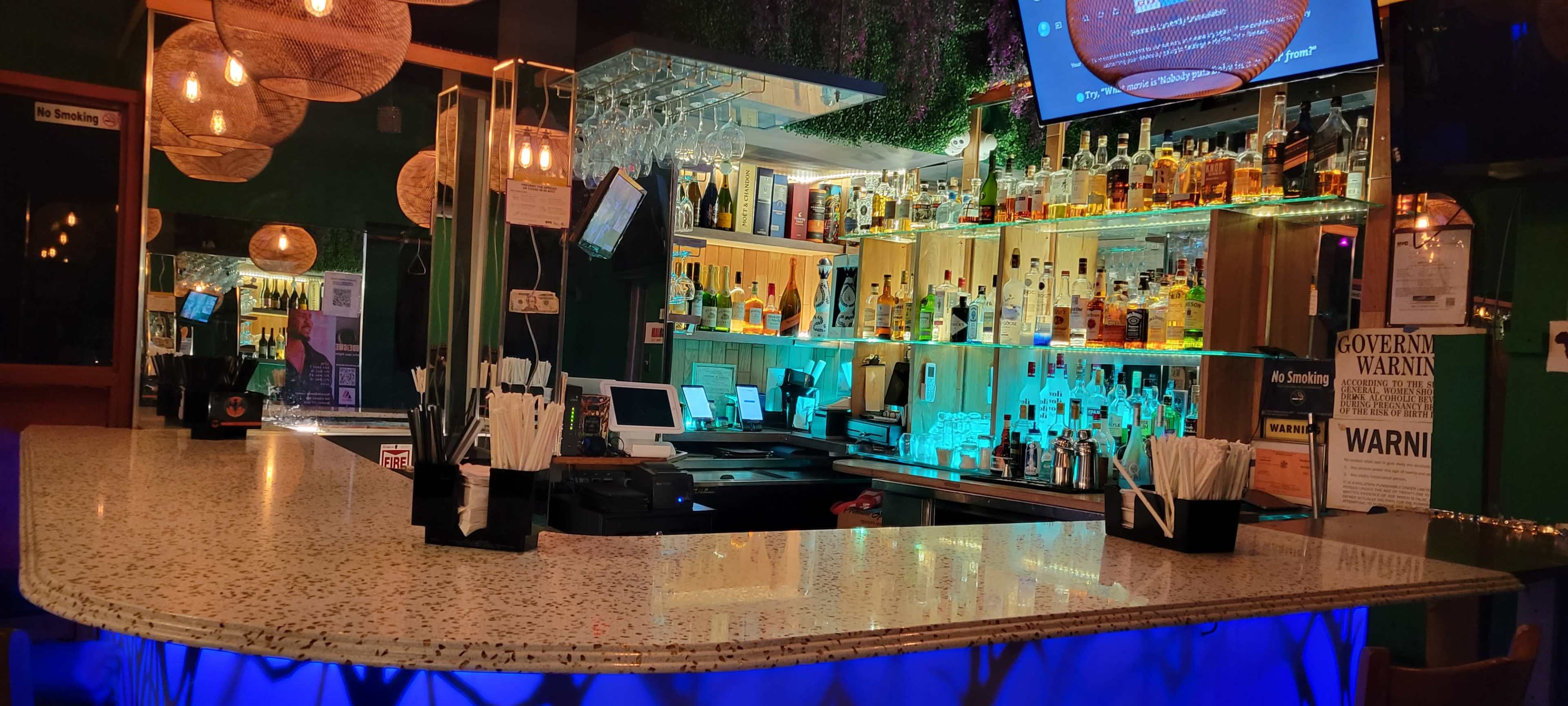 The image shows a well-lit bar area featuring a long countertop, backlit shelves stocked with various bottles, and several hanging light fixtures.