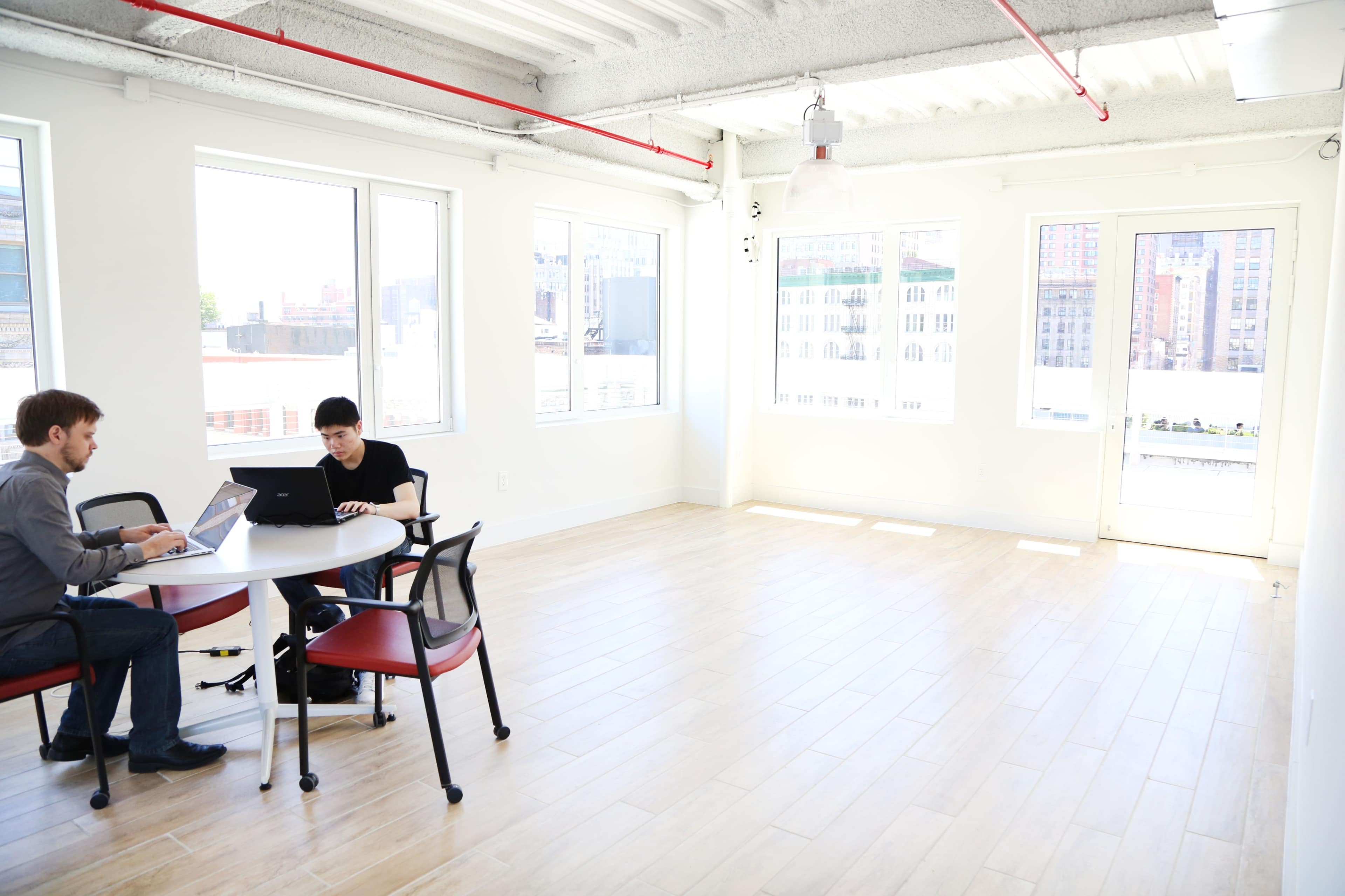 Two people are working on laptops at a round table in a bright, empty office space with large windows.