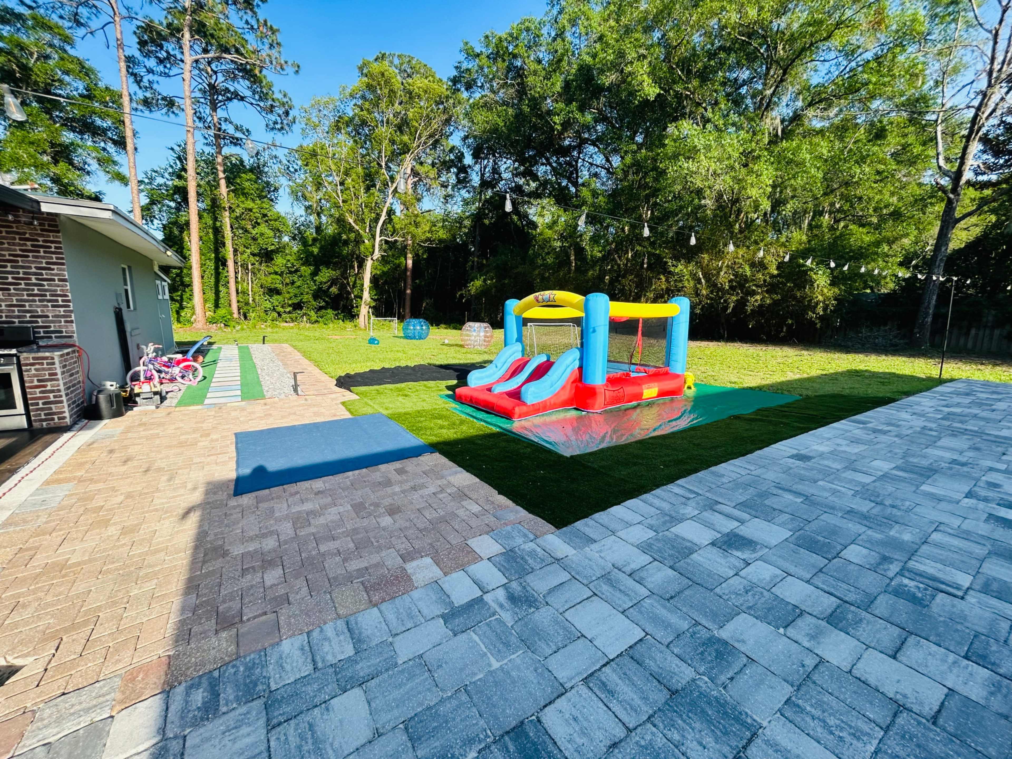 The image shows a backyard with a bounce house, a paved area, and playground equipment surrounded by trees.