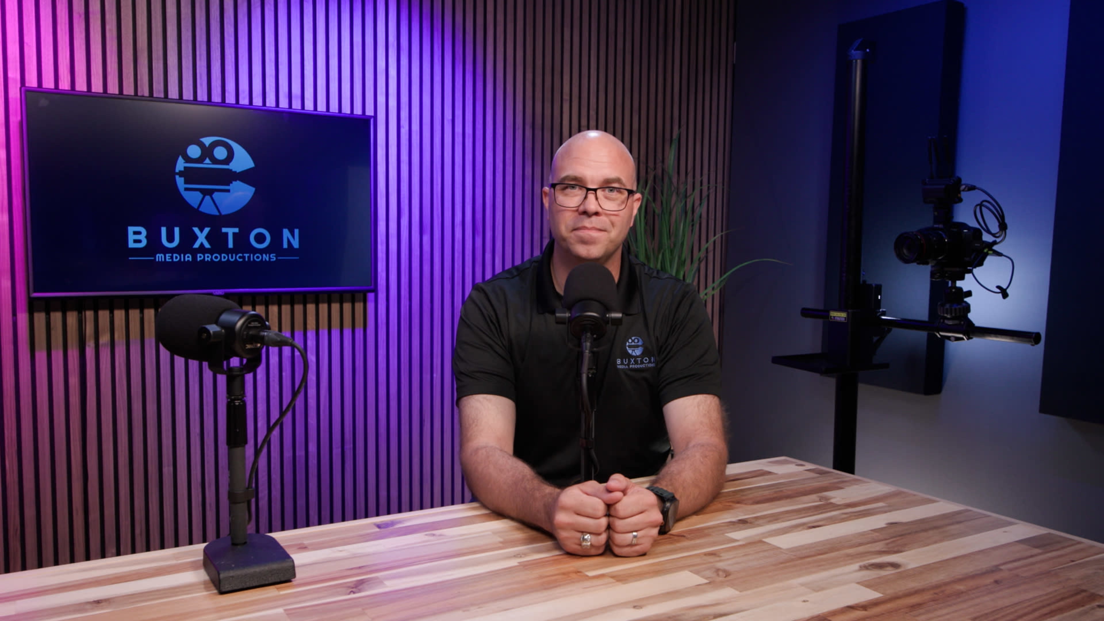 A man sits at a wooden table in a studio with microphones and a camera, facing a wall displaying the "Buxton Media Productions" logo.