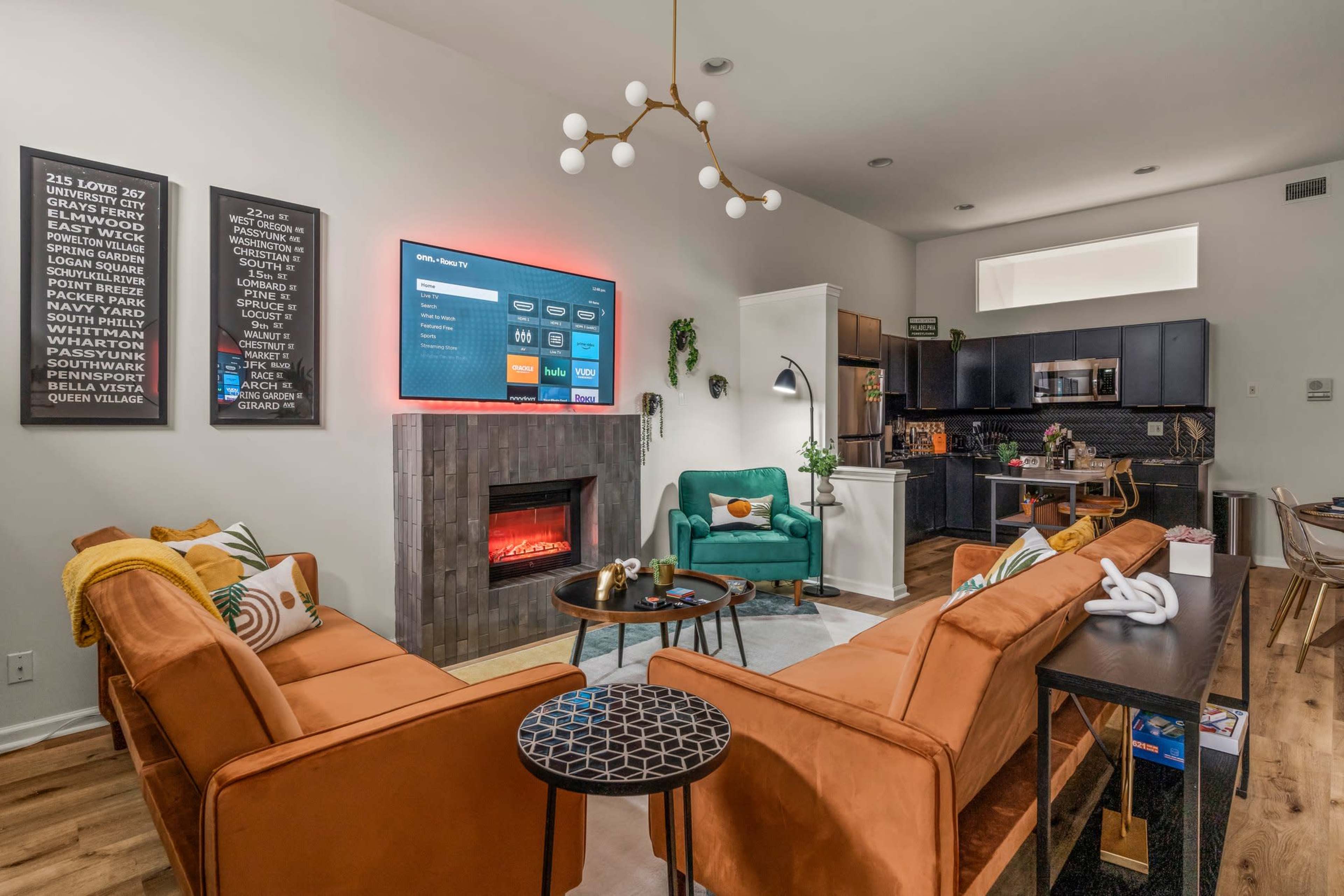A modern living room featuring orange sofas, a mounted television, a fireplace, and a kitchenette in the background.