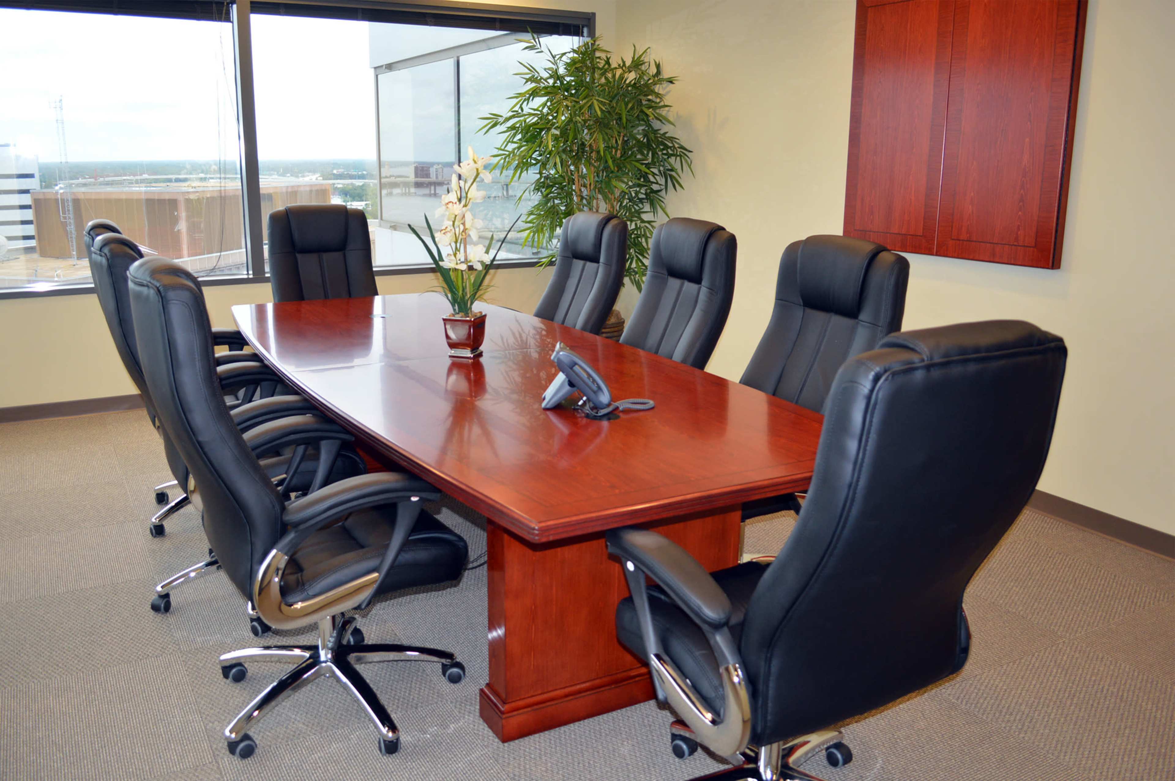 The image shows a conference room with a large wooden table surrounded by six black leather office chairs, a phone, and a potted plant near a window.