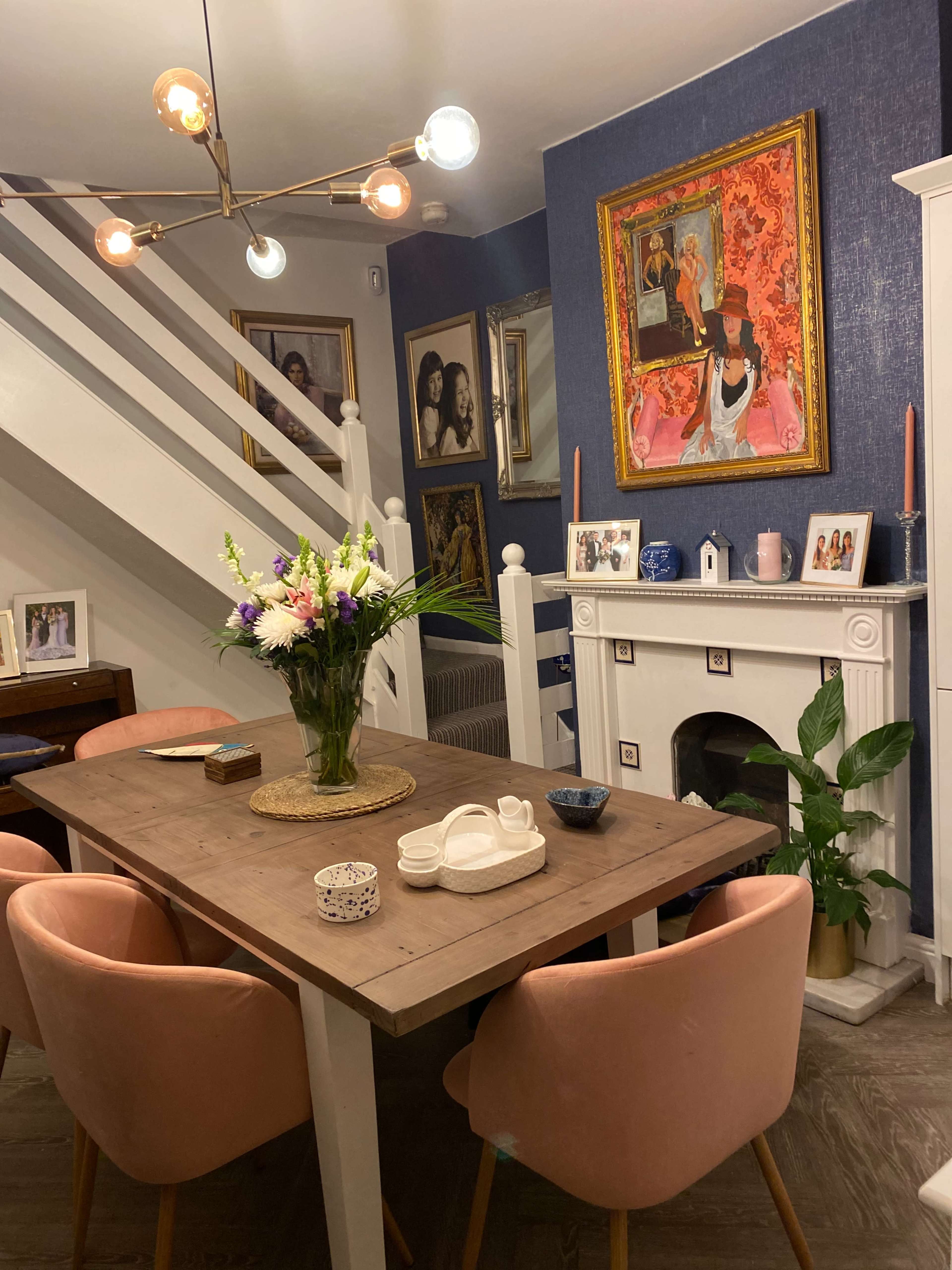 A dining area with a wooden table surrounded by pink chairs, a floral centerpiece, and paintings on the wall, including a prominent artwork above a white fireplace.