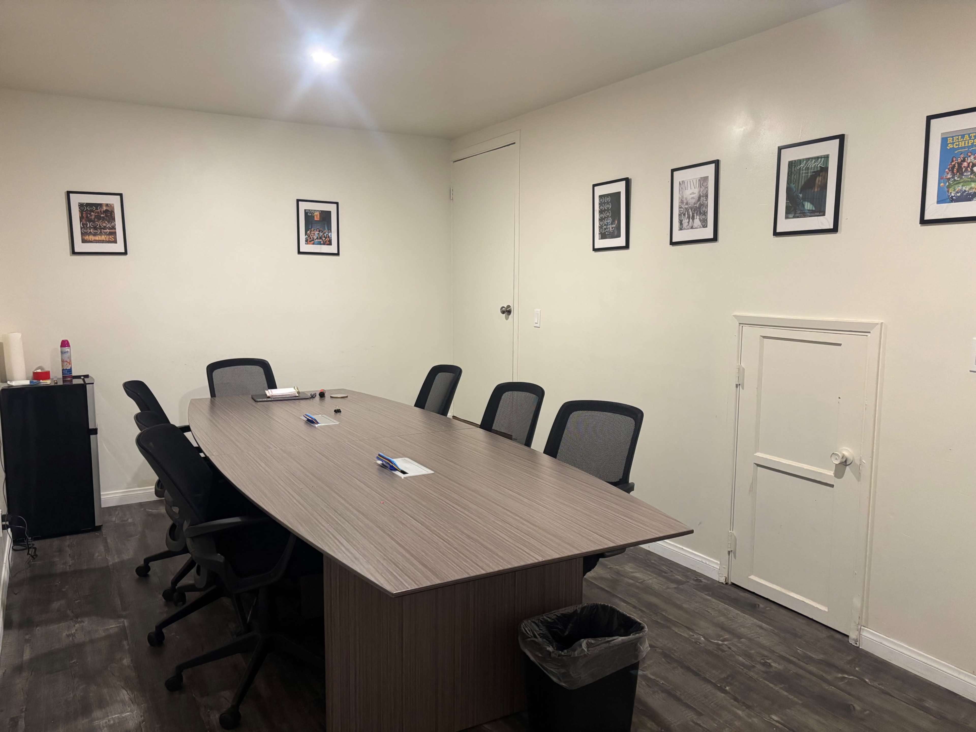 The image shows a conference room with a long wooden table surrounded by several black chairs, and framed photographs displayed on the white walls.