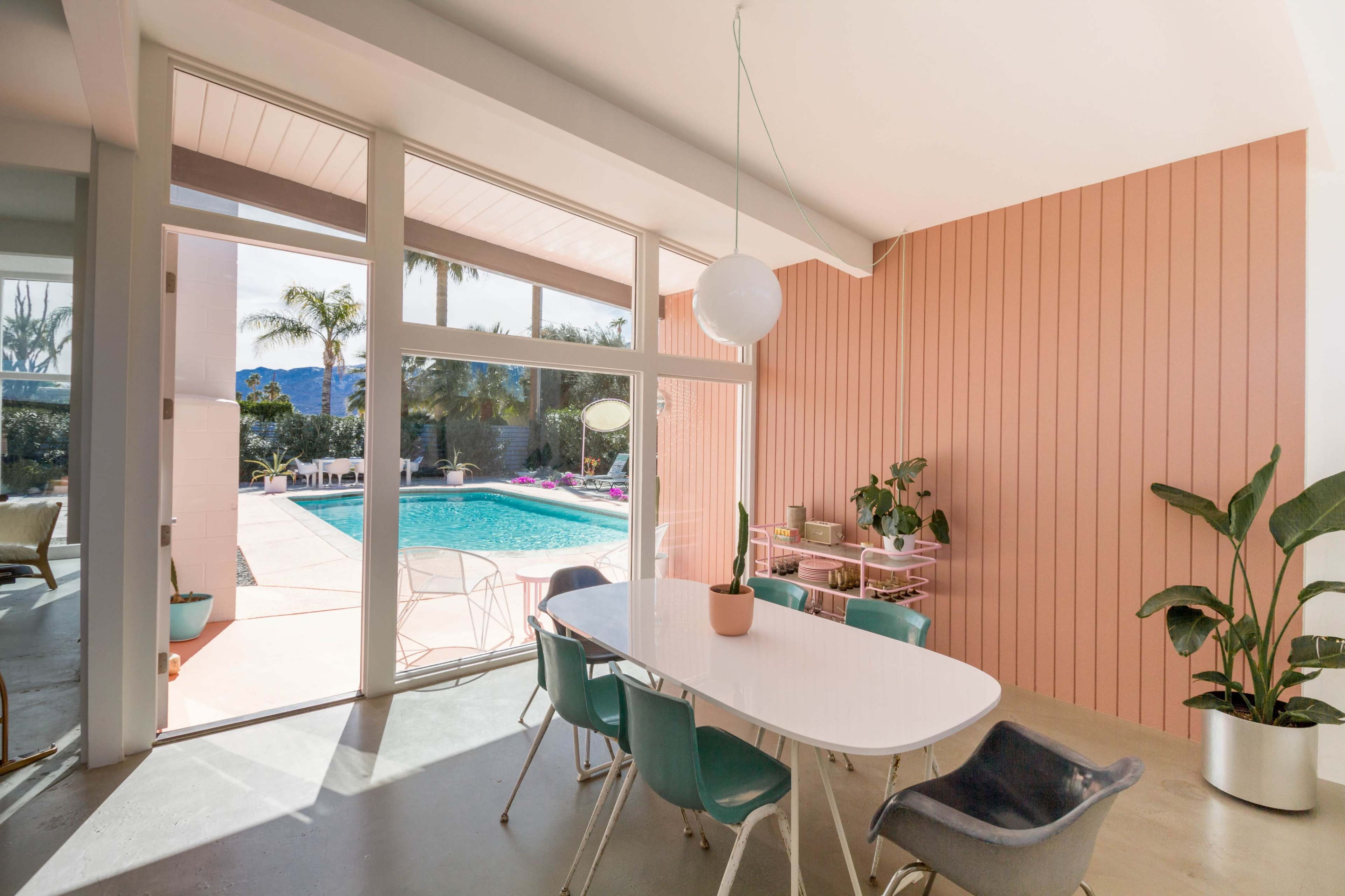 The image shows a dining area with a white table and colorful chairs, open to a poolside view framed by large windows and a pink wall.