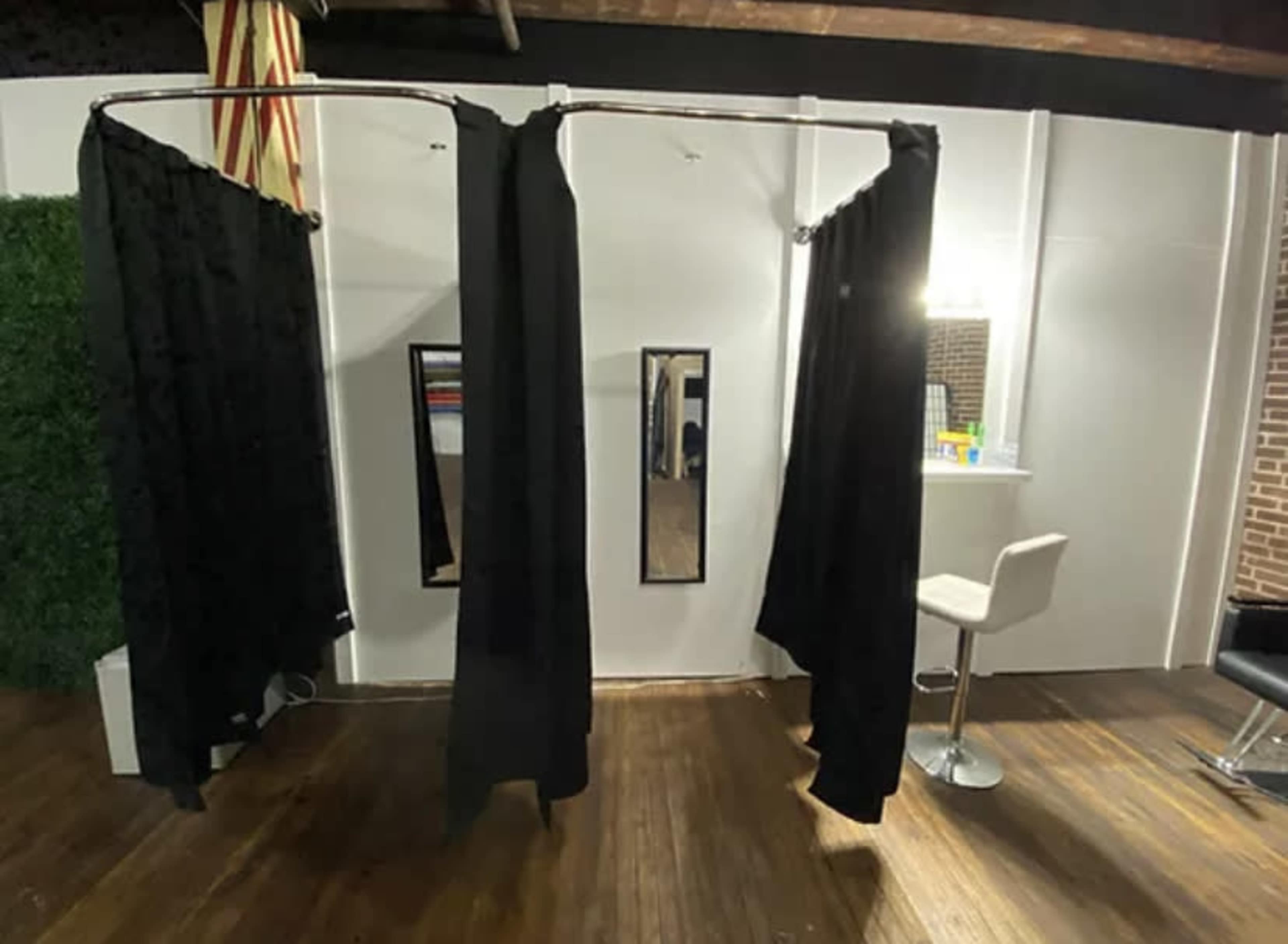 The image shows a room divided by black privacy curtains, featuring a mirror and a chair near a brightly lit counter.