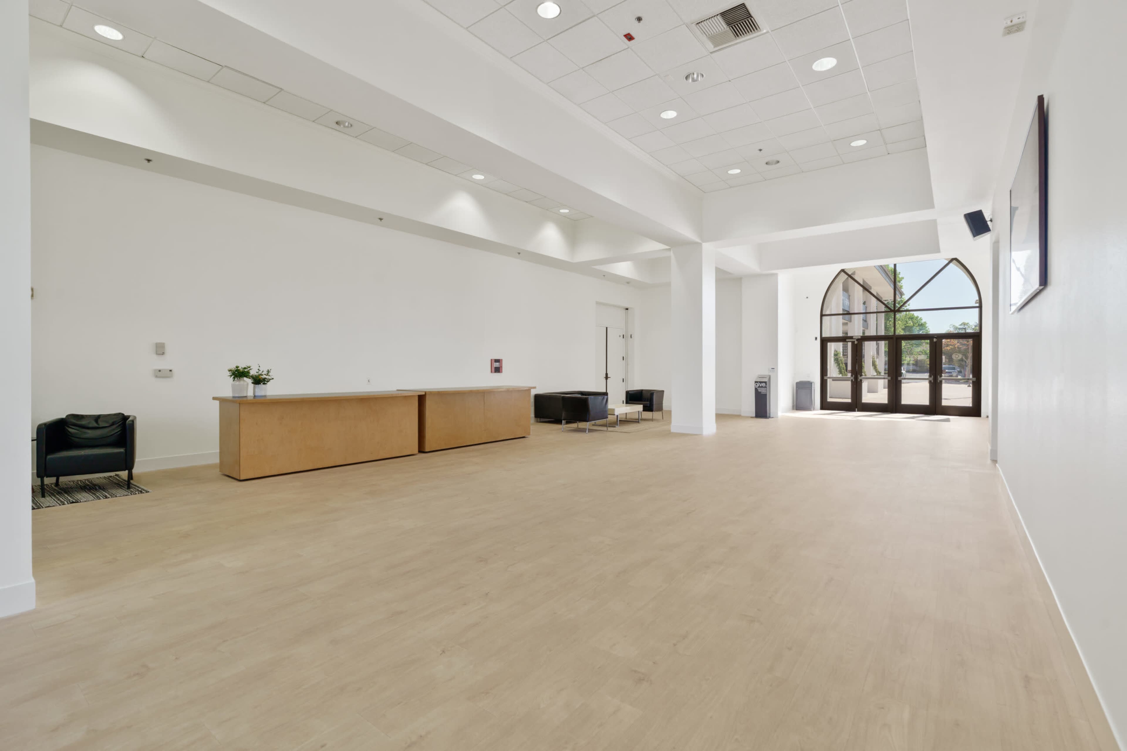 The image shows a spacious, empty interior lobby with a wooden reception desk, black seating, and large arched doors leading outside.
