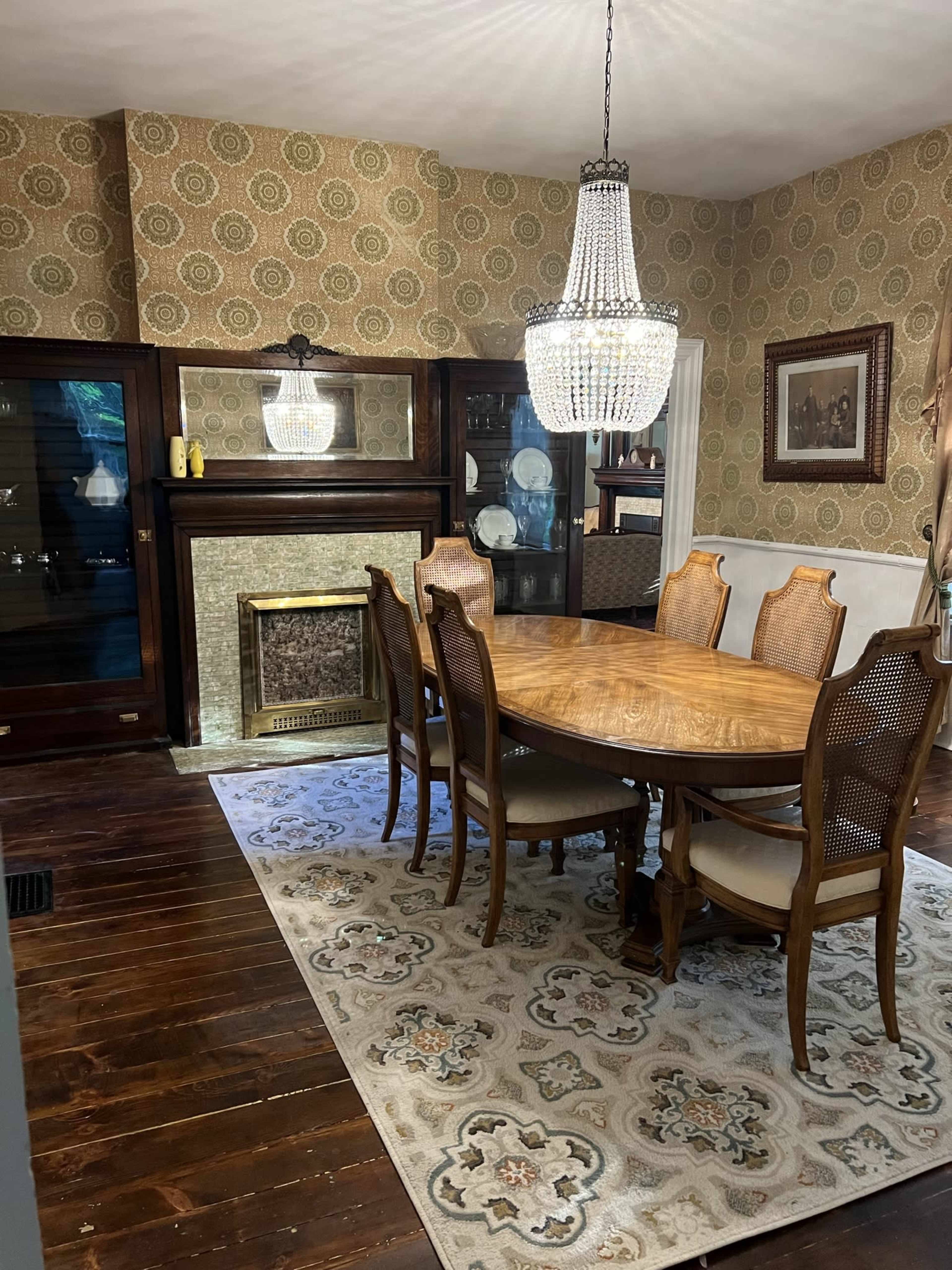 A dining room features a wooden table surrounded by six chairs, a chandelier overhead, and a fireplace adorned with patterned wallpaper and a decorative rug.