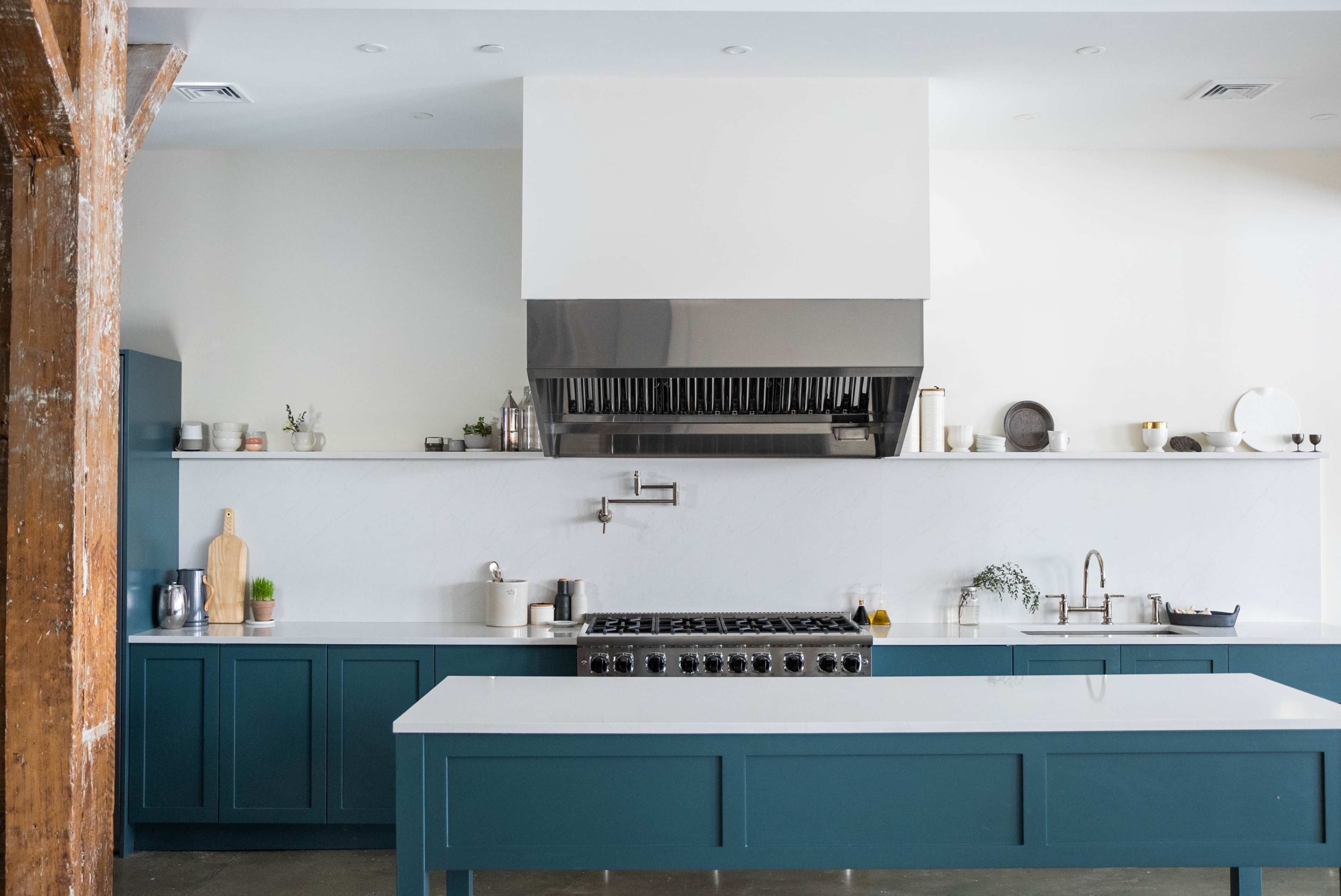 A modern kitchen features a large island with a stove, stainless steel hood, and minimalist cabinetry in teal and white.