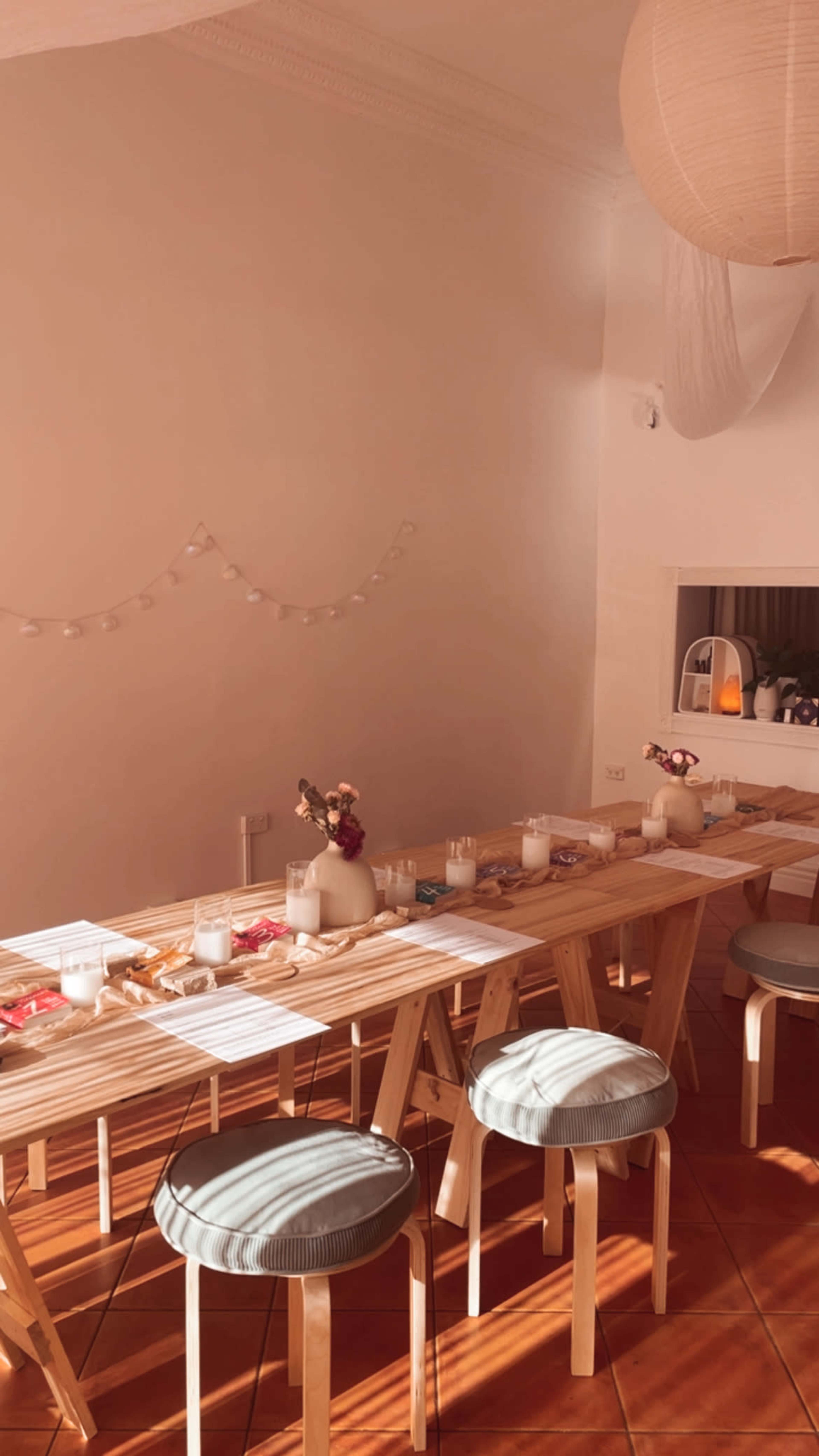 A long wooden table is set with candles and decorative items, surrounded by stools in a softly lit, minimalist interior.