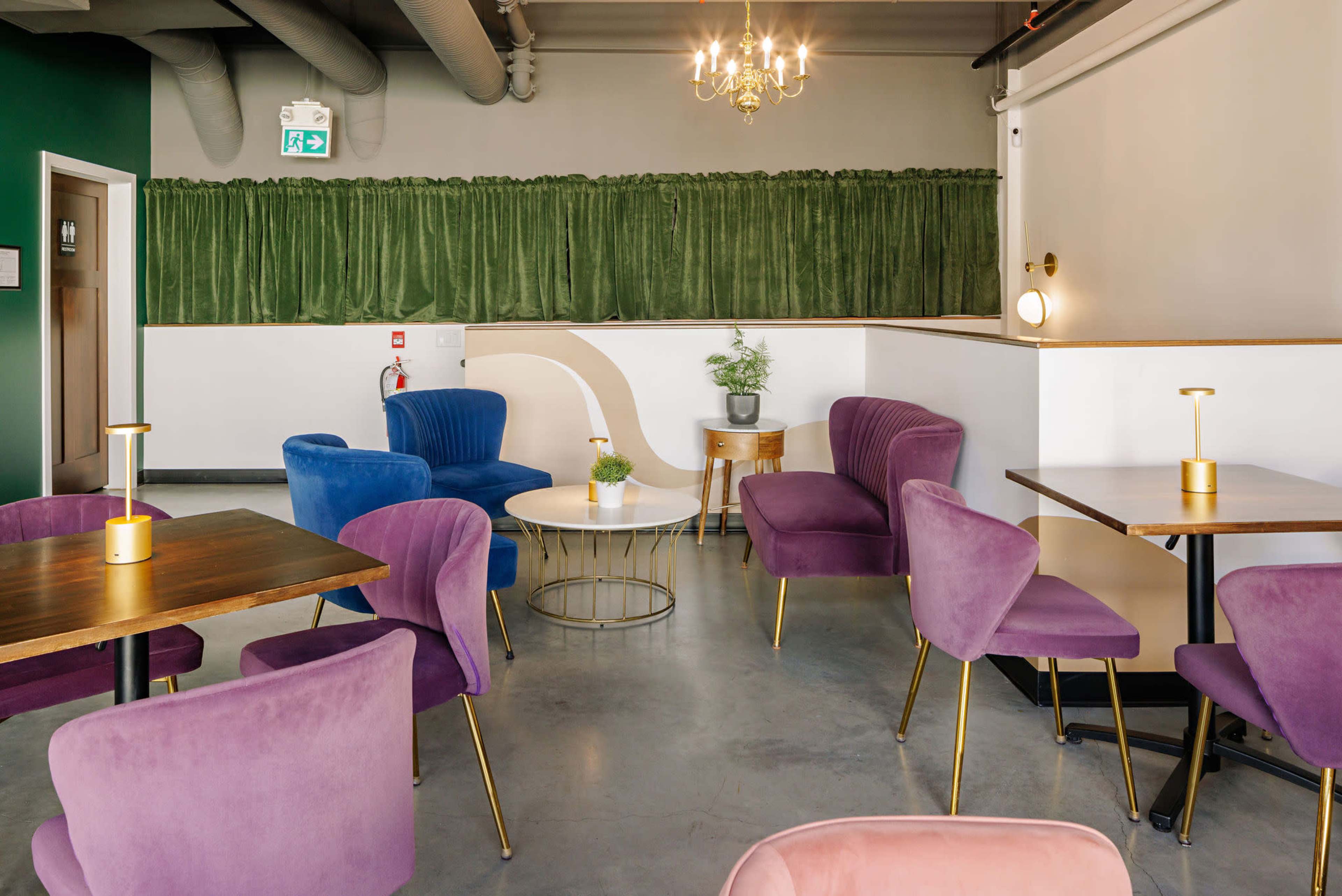 The image shows a modern interior of a café featuring purple and blue upholstered chairs, wooden tables, and green drapery along a wall.