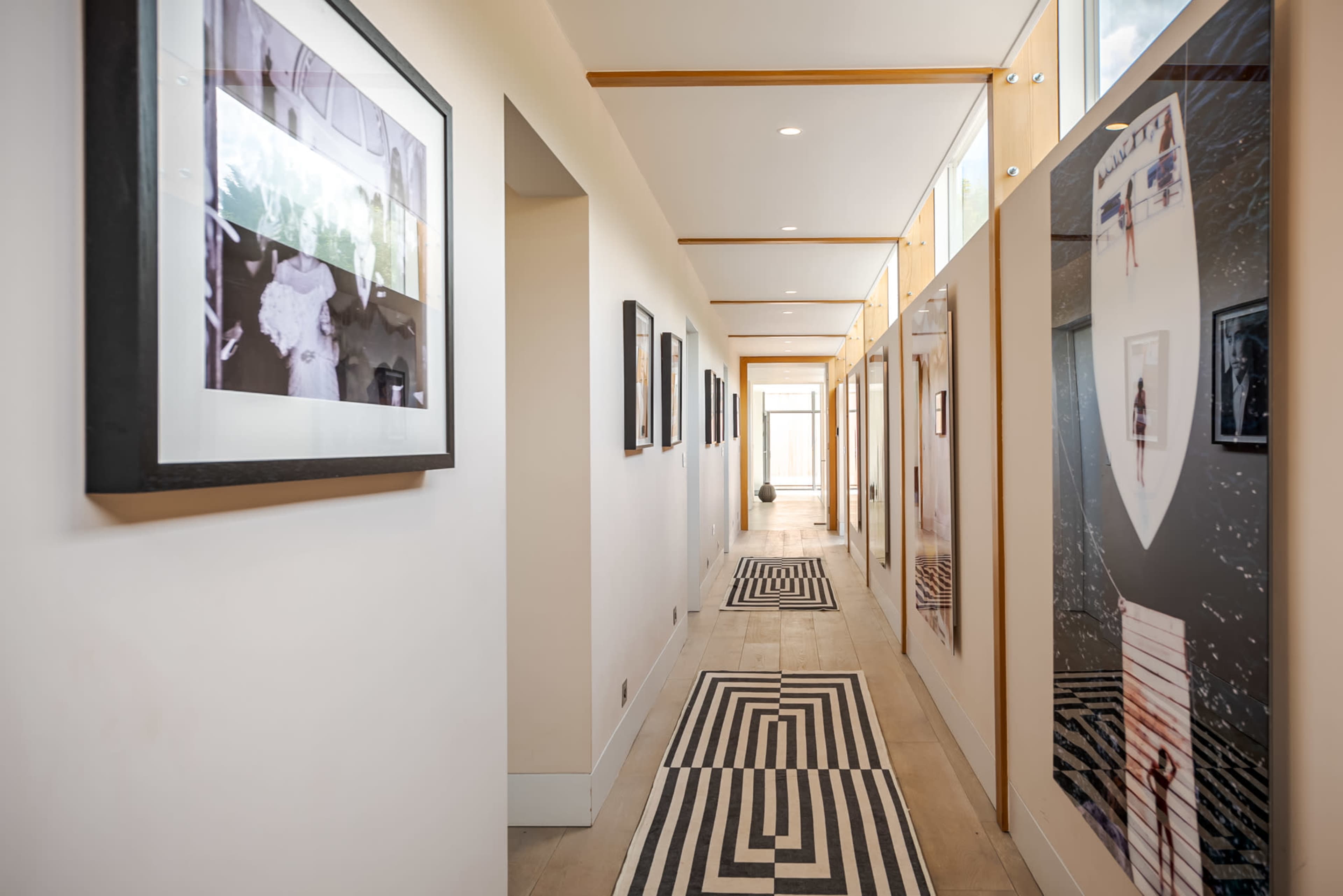 The image shows a modern hallway lined with framed photographs on both sides, featuring a striped black-and-white rug on the floor.