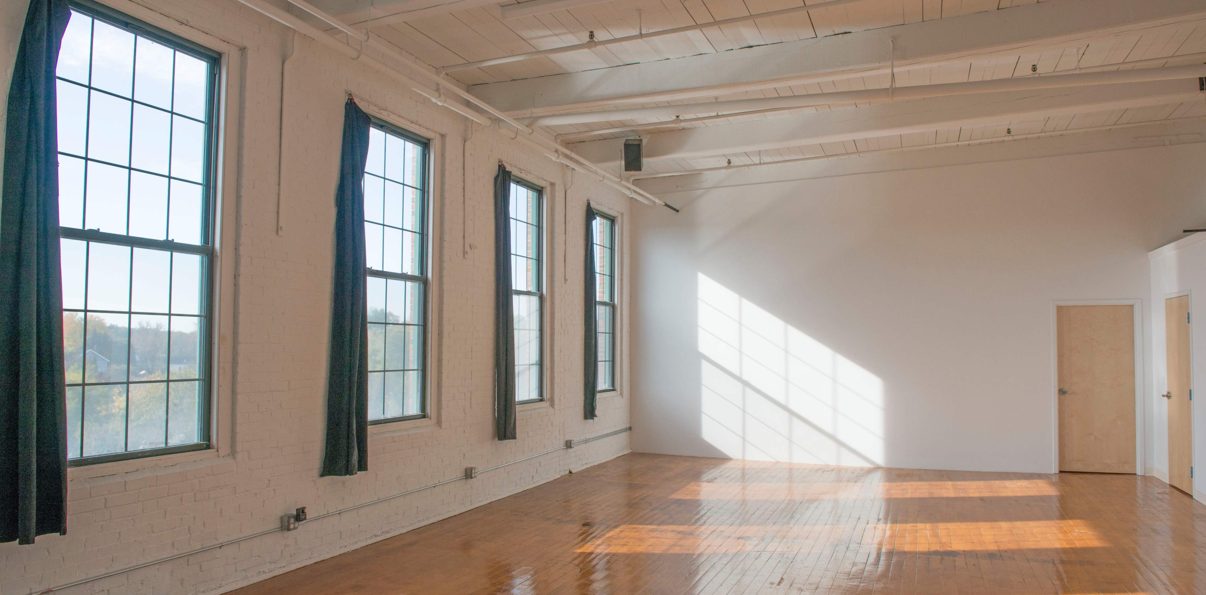 A bright, empty room features large windows and wooden flooring, with light casting shadows on the wall.