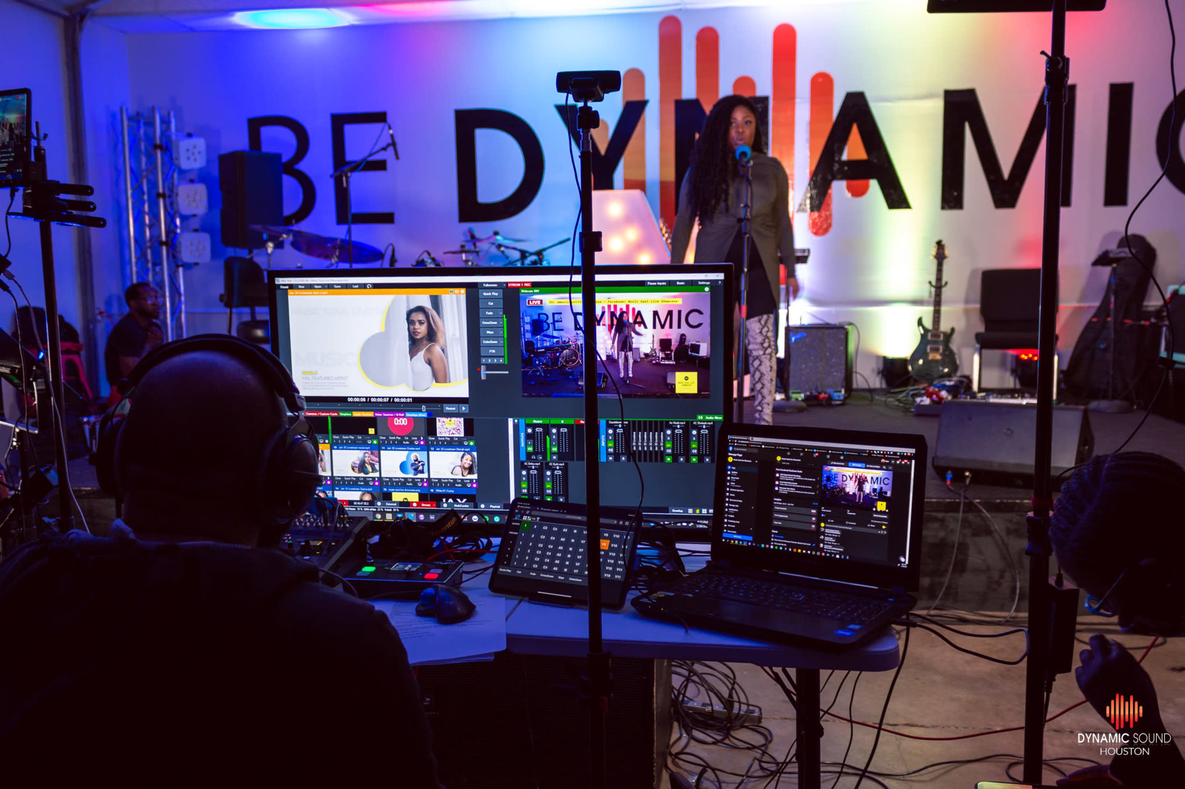 A performer sings on stage in front of a large "BE DYNAMIC" sign, while a sound technician operates multiple screens and audio equipment.