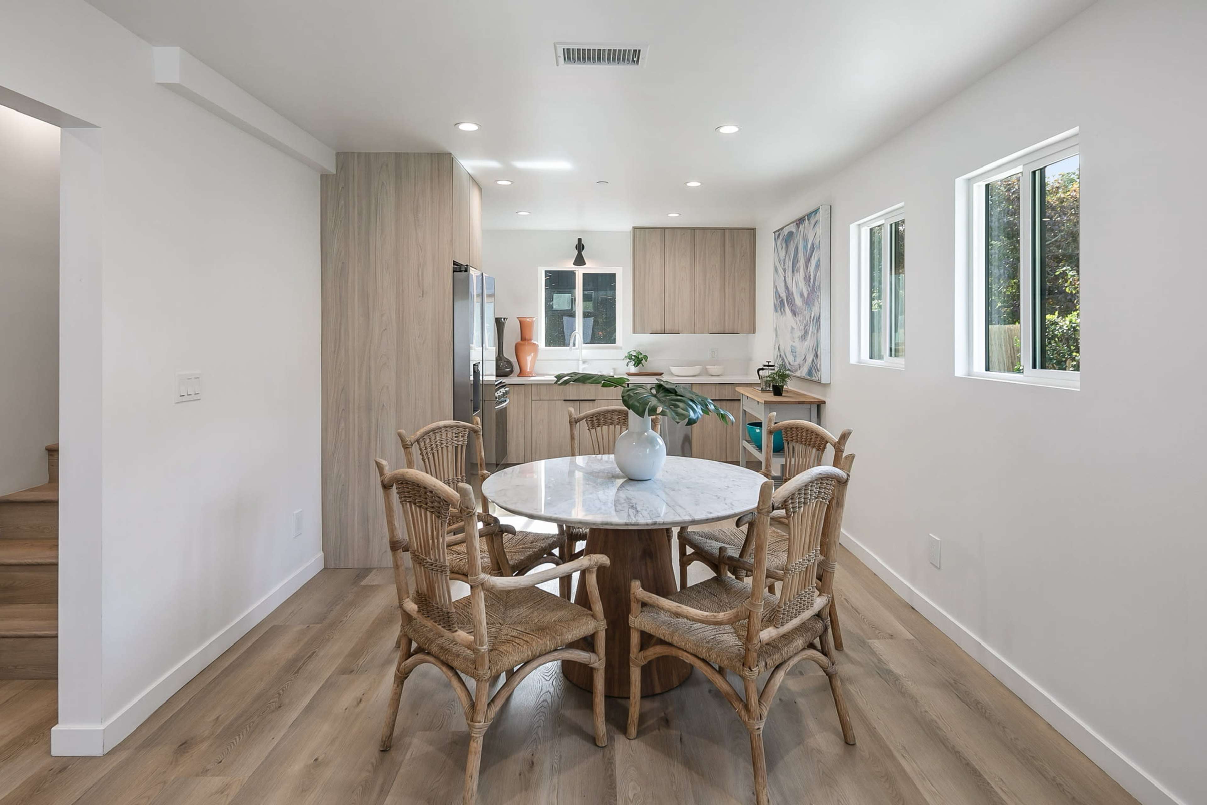 A modern kitchen with a round marble dining table surrounded by wicker chairs, set against a backdrop of wooden cabinets and large windows.