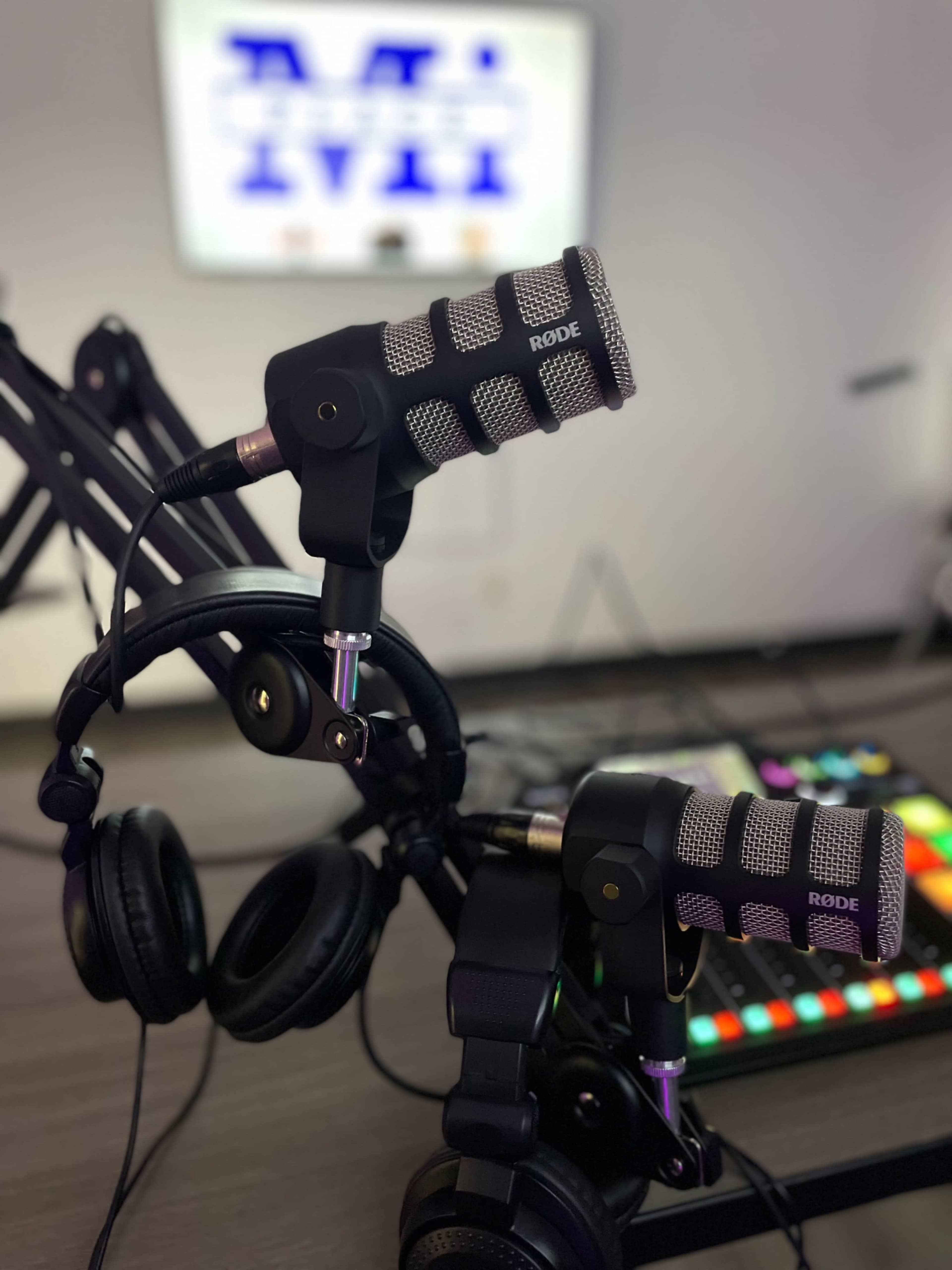 Two Rode microphones are mounted on adjustable arms beside a pair of black headphones and a colorful control panel in a recording studio.