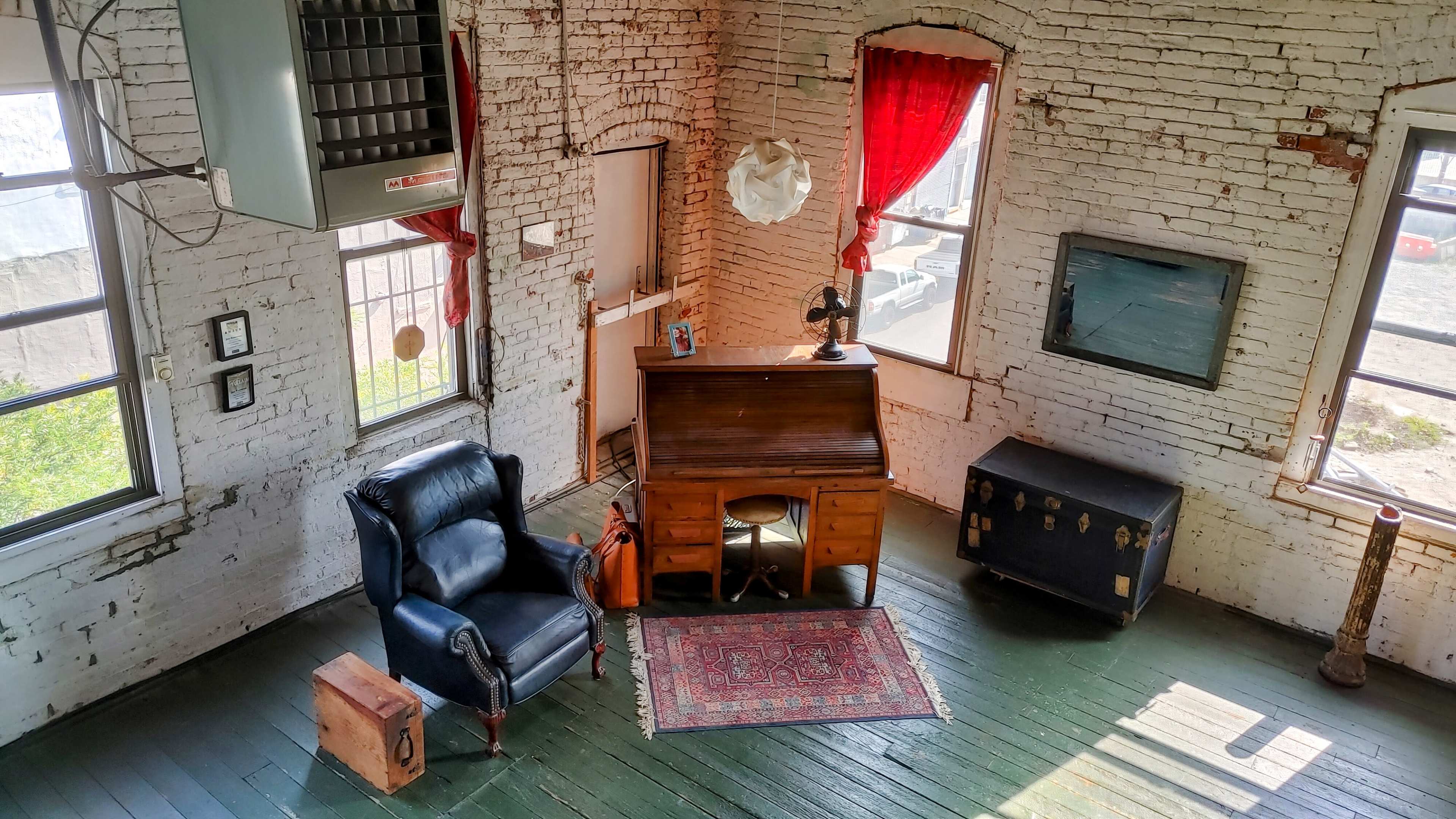 The image shows a room with exposed brick walls, a vintage writing desk, a black leather chair, and red curtains framing two windows.