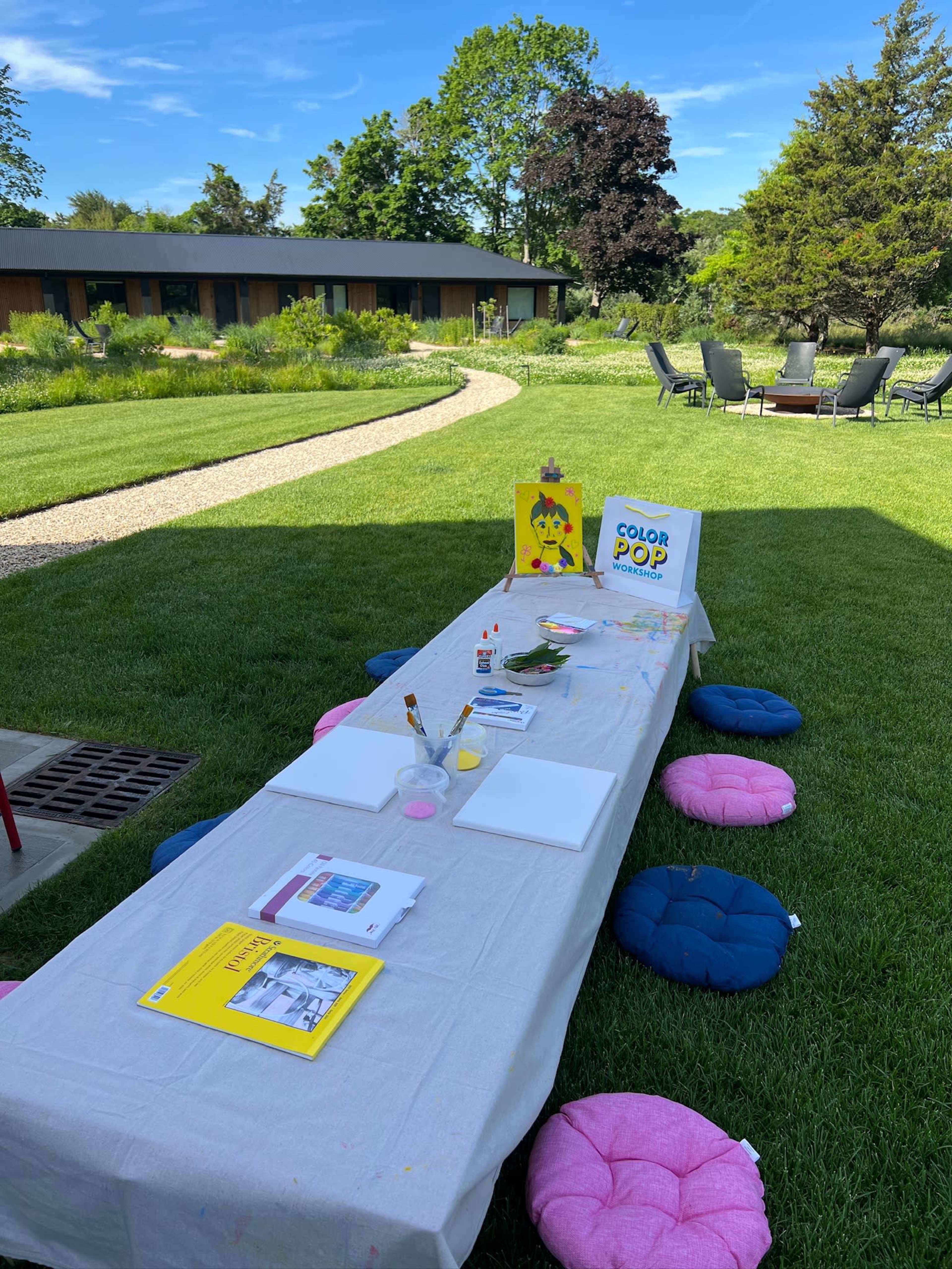 A long table covered with a gray tablecloth is set up in a green yard for a painting activity, surrounded by colorful cushions and a background of trees and a building.