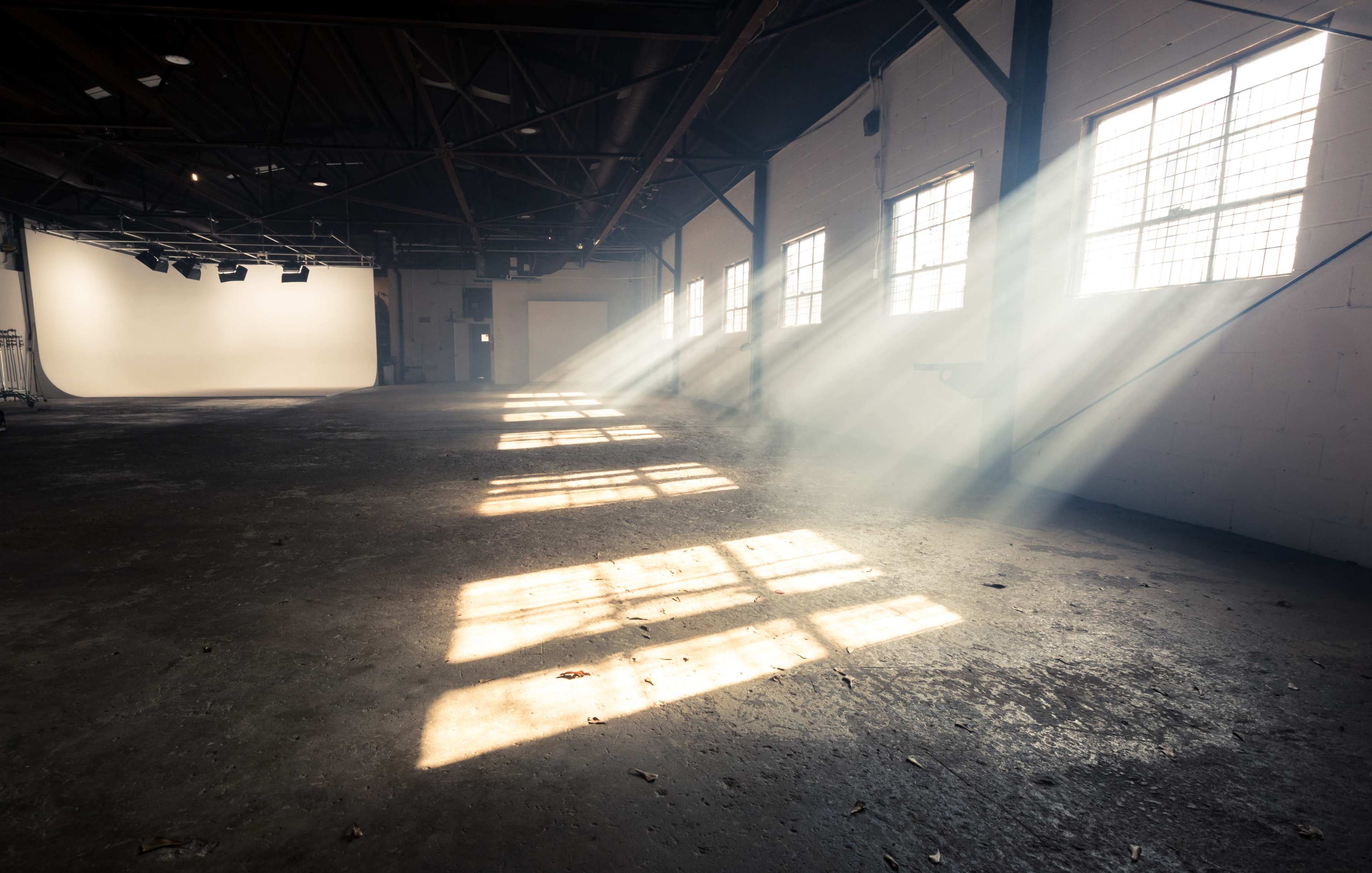 Sunlight streams through the windows of a spacious, empty warehouse, casting distinct shadows on the concrete floor.