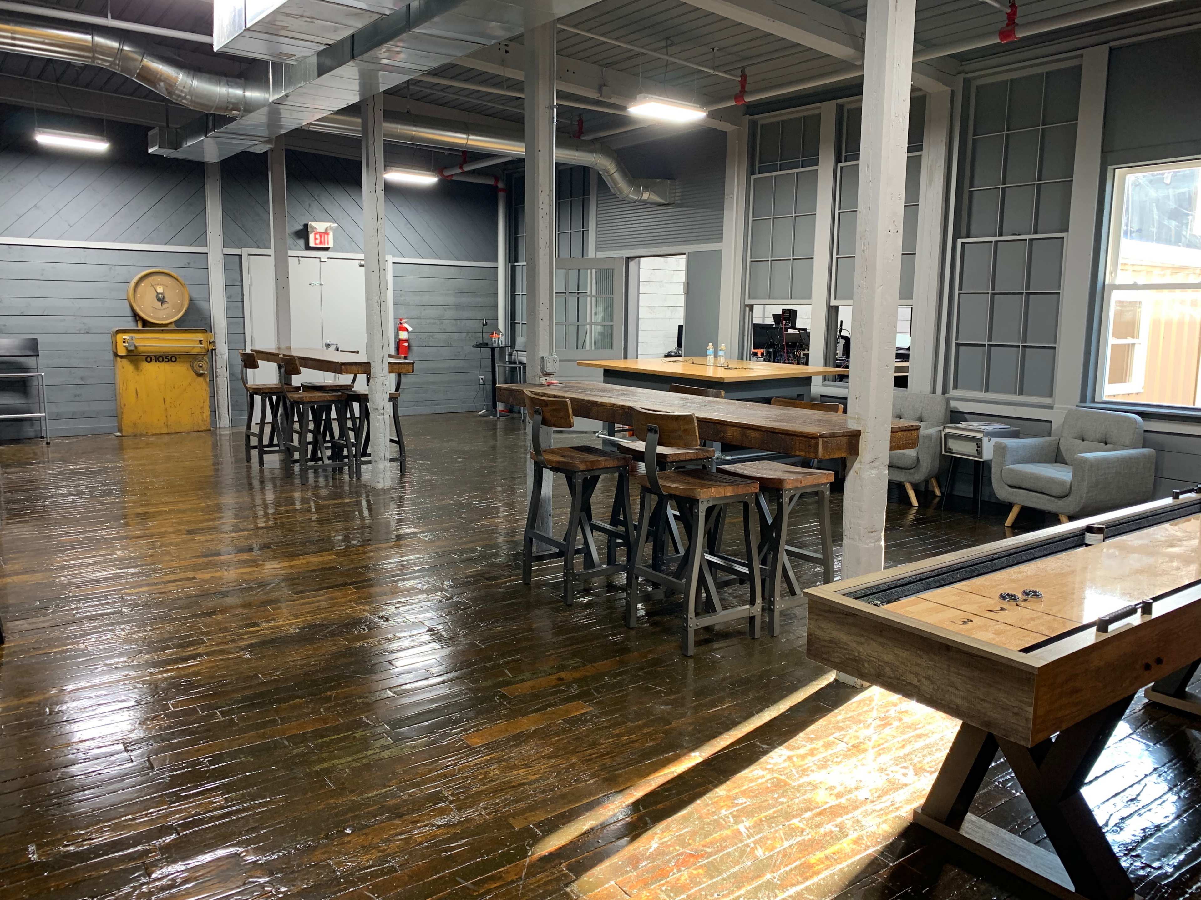 The image shows a spacious interior with polished wood floors, several wooden tables and chairs, a bar counter, and large windows allowing natural light.