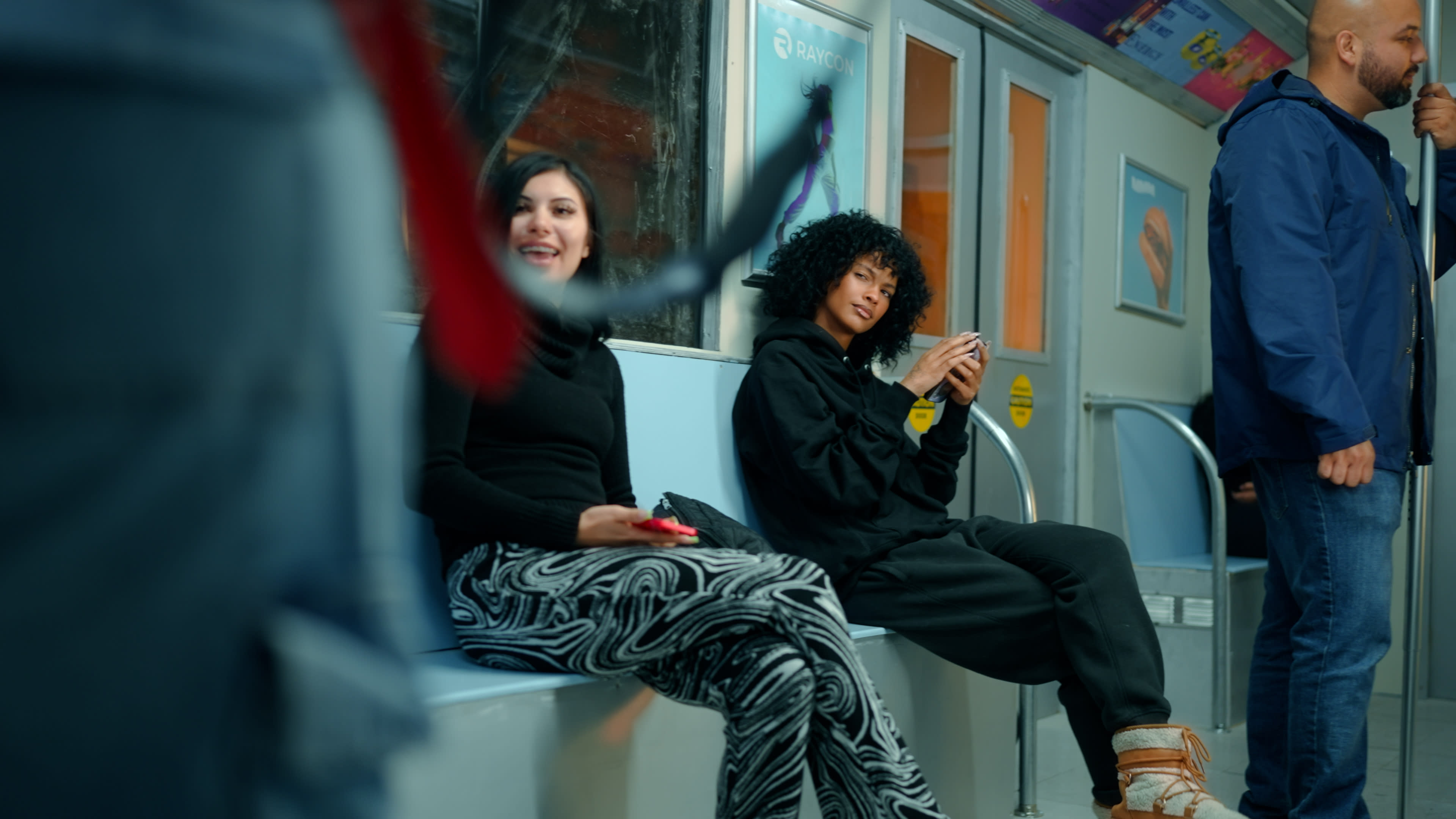 Two individuals sit on a subway bench, one smiling while the other looks at their phone, as a few people stand nearby.