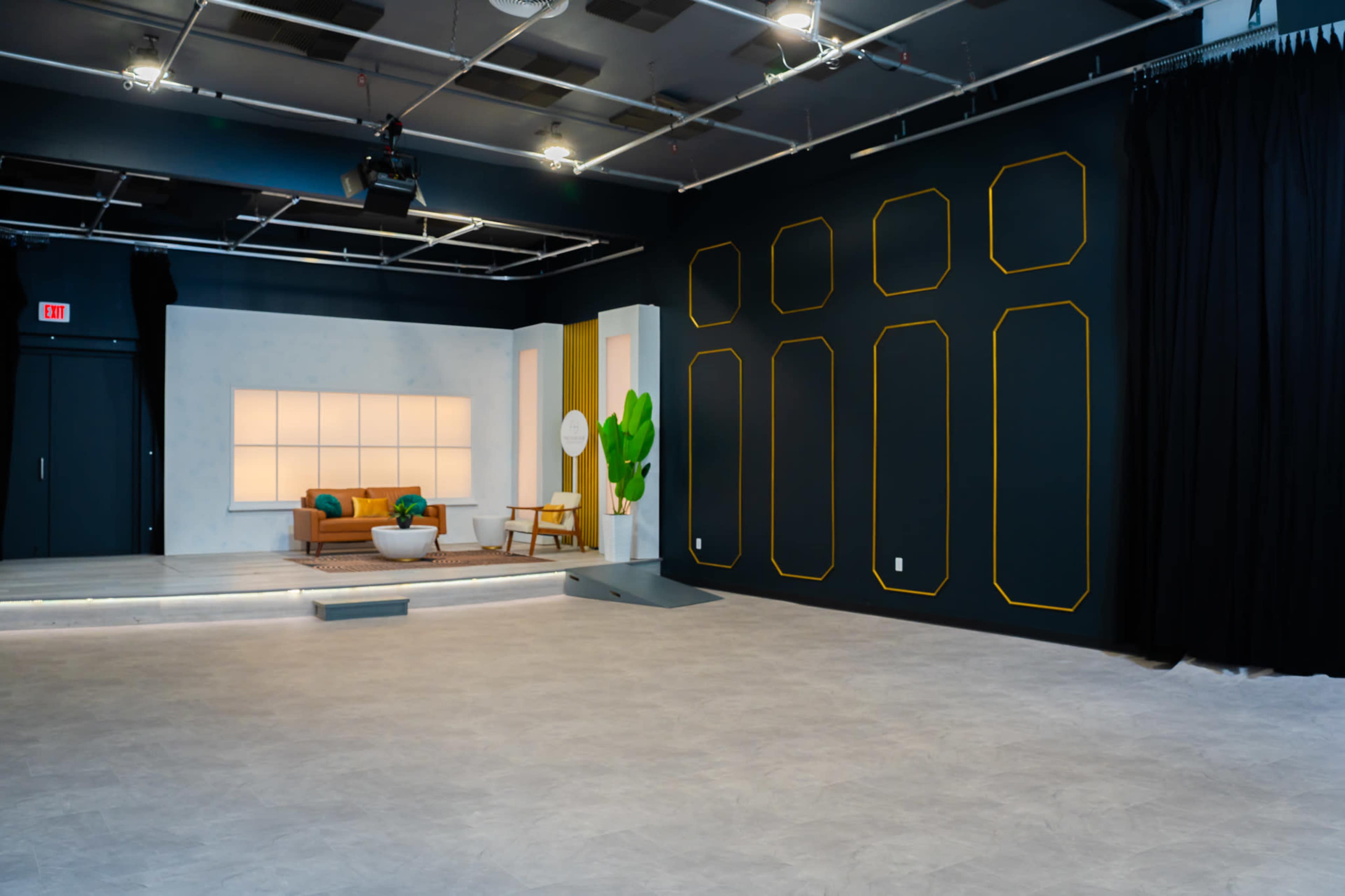 The image shows a modern studio space with a light-colored seating area against a backdrop of dark walls featuring geometric outlines and a wall with black curtains.