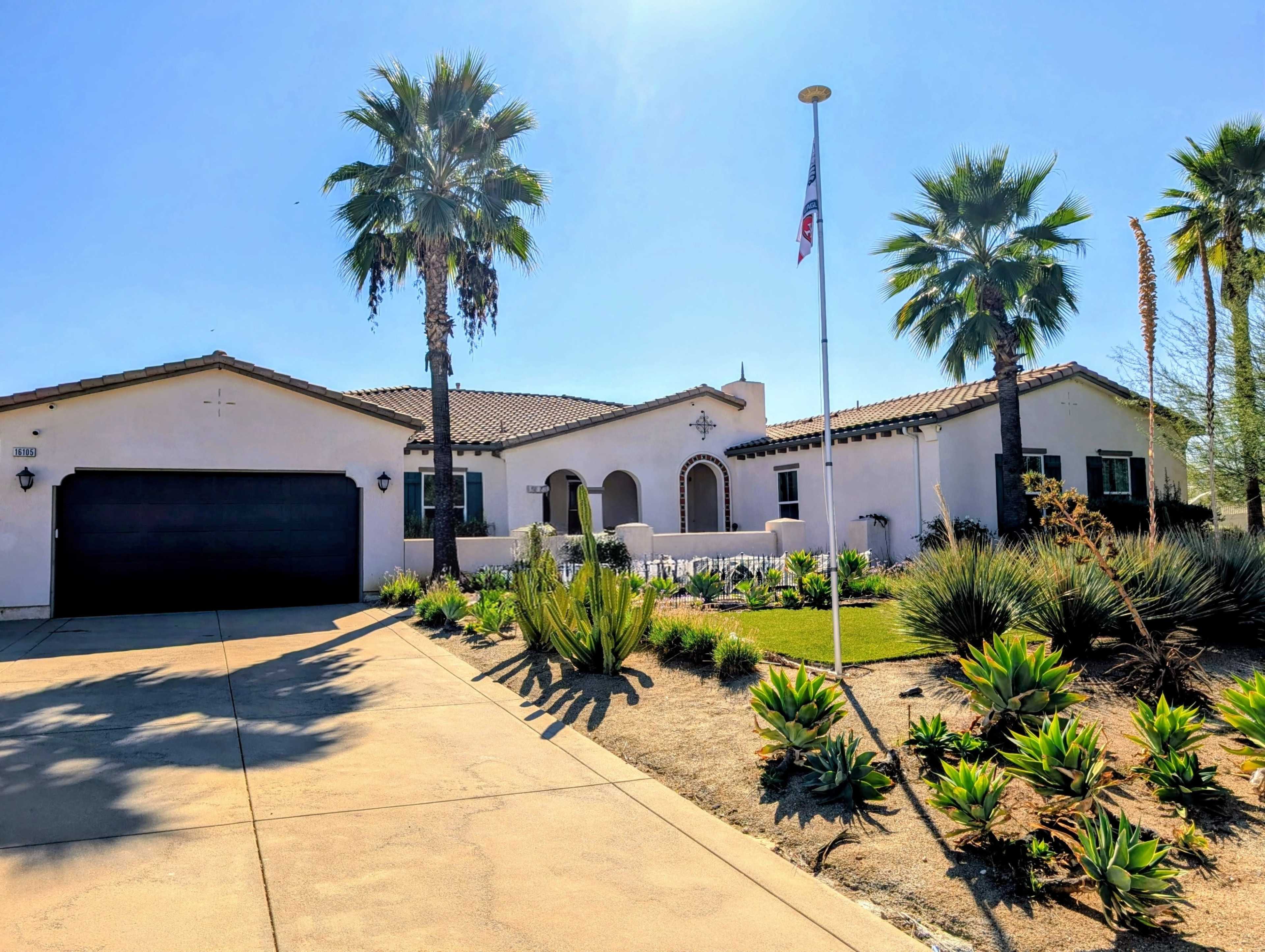 A single-story house with a black garage door, surrounded by palm trees and landscaped with various plants.