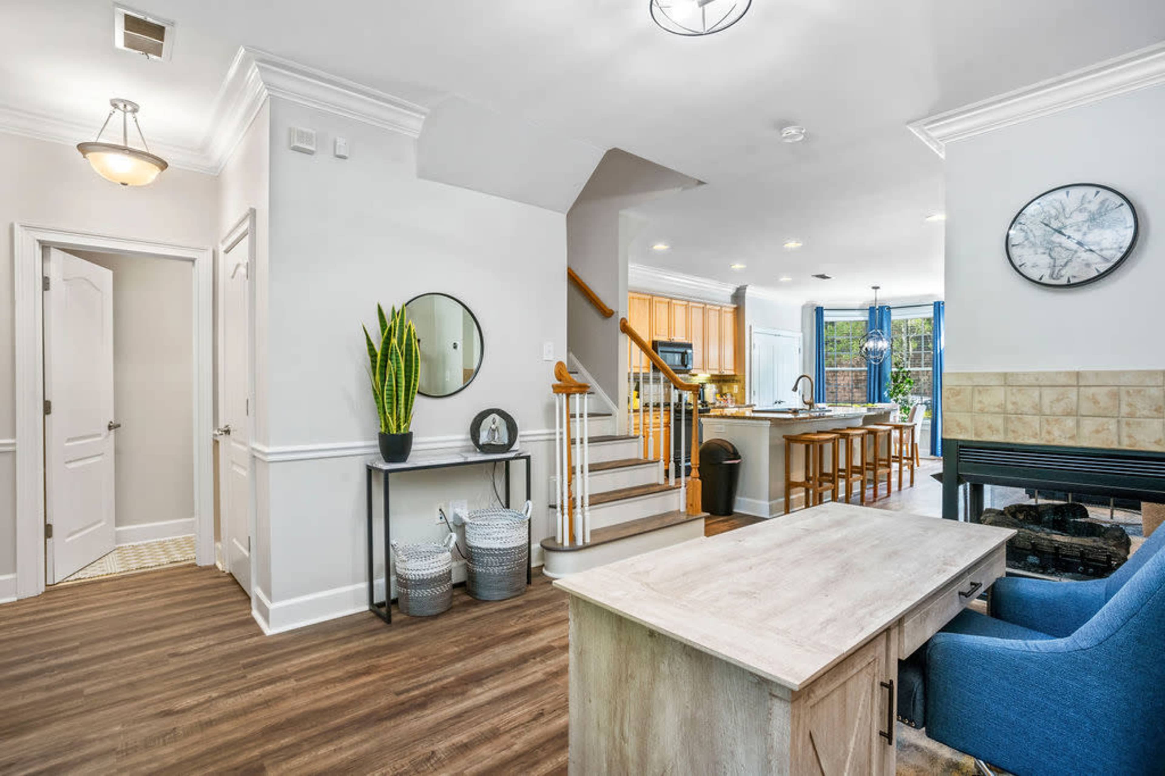 The image shows a bright, open-entryway leading to a staircase, with a living area and kitchen visible in the background.