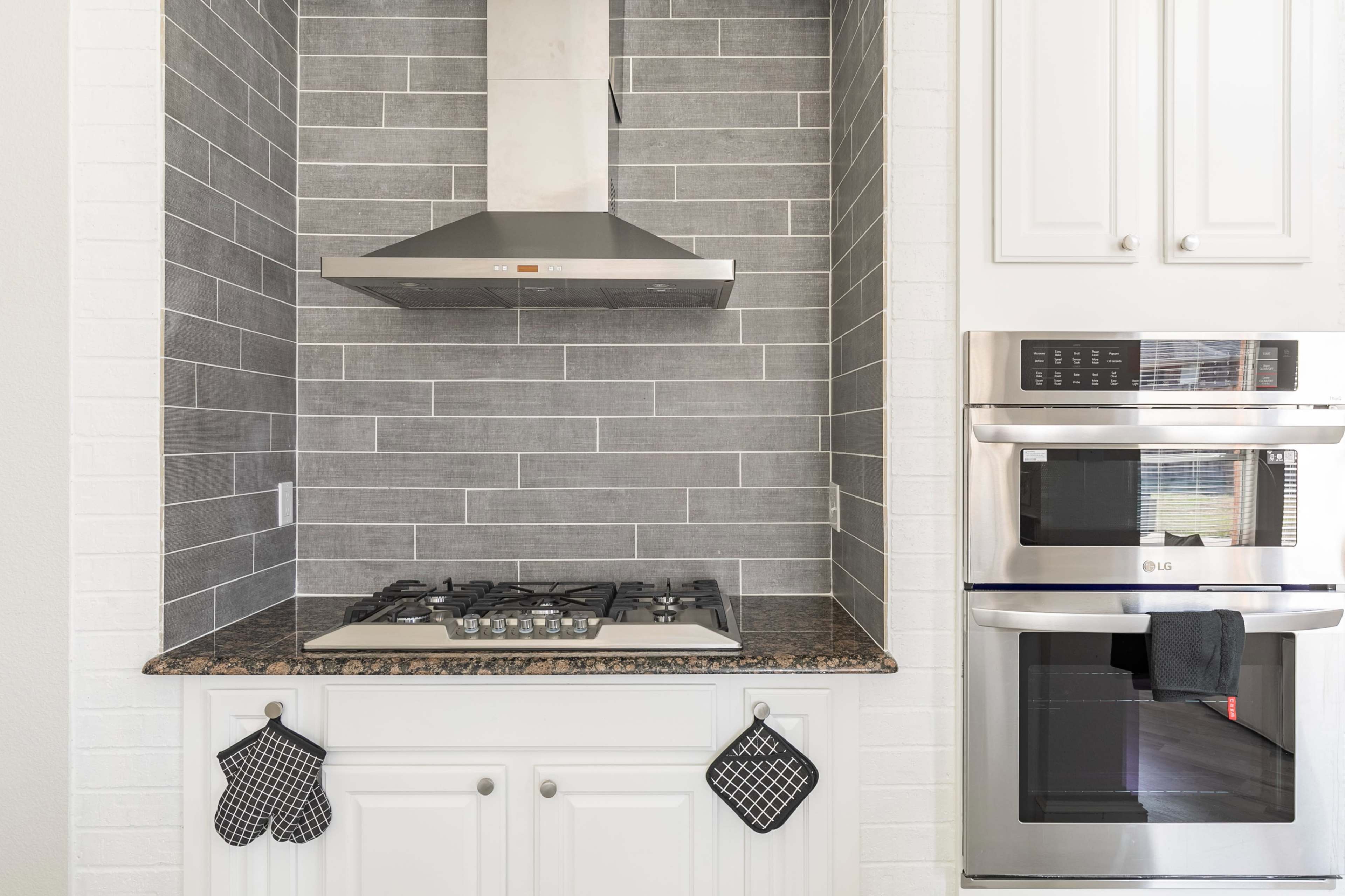 A kitchen area featuring a gas stove, a stainless steel hood, and a wall oven, with a gray tiled backsplash and dark granite countertop.