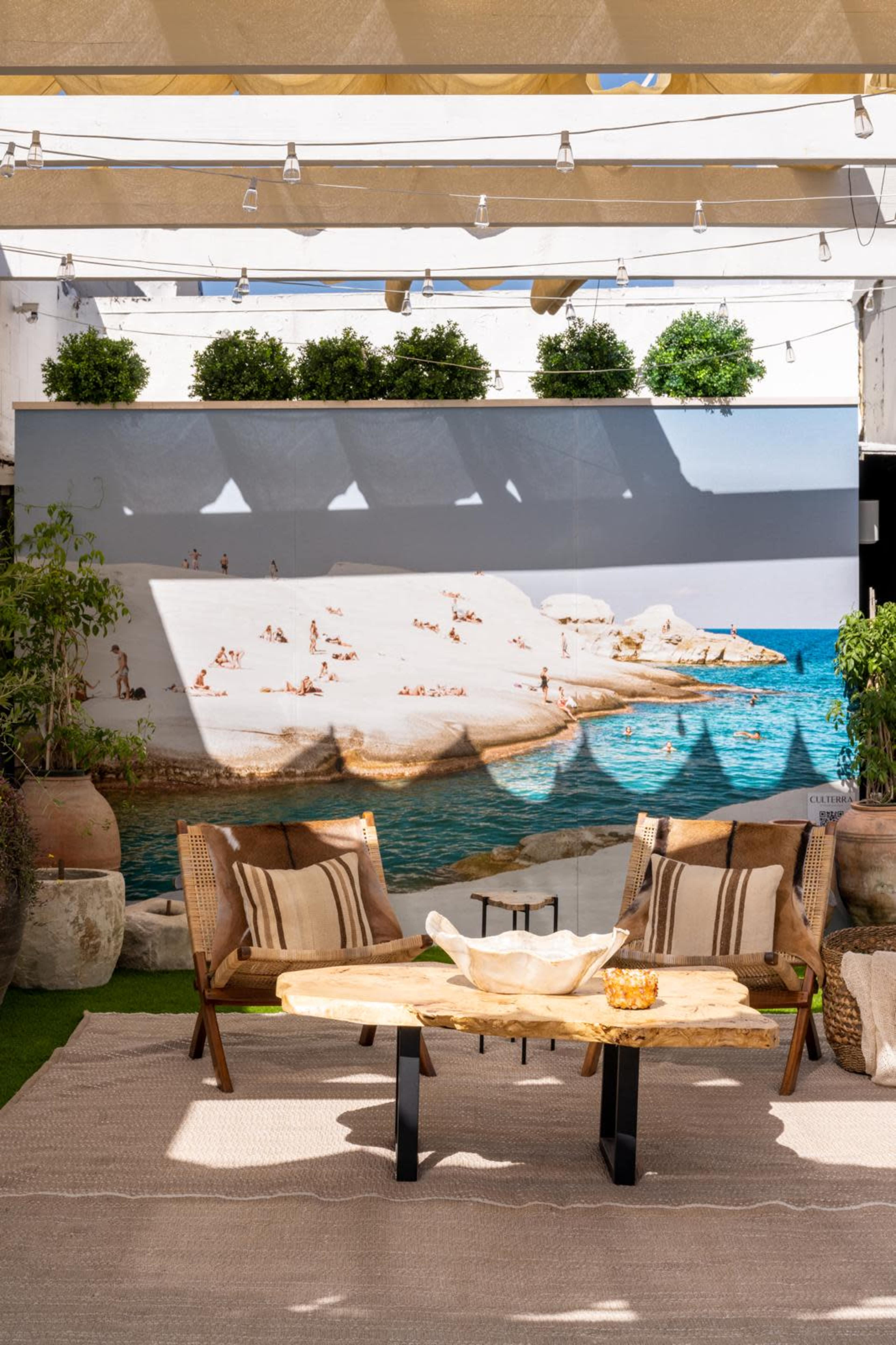 The image shows an outdoor seating area with two chairs and a wooden table, set against a large mural depicting a beach scene with people sunbathing by the water.