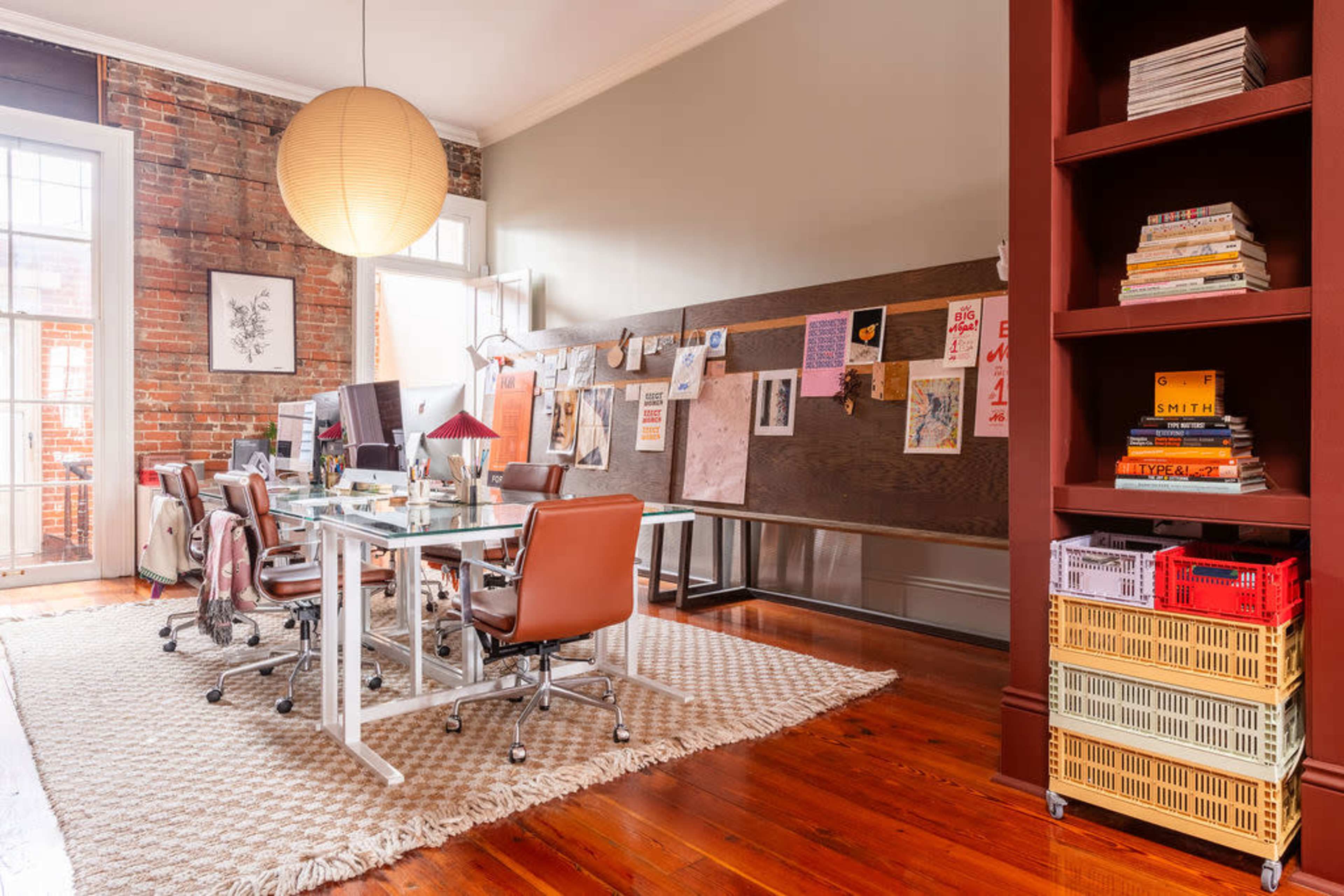 A modern office space features several glass desks with ergonomic chairs, a wooden accent wall displaying various artworks, and a red bookshelf filled with colorful baskets and books.