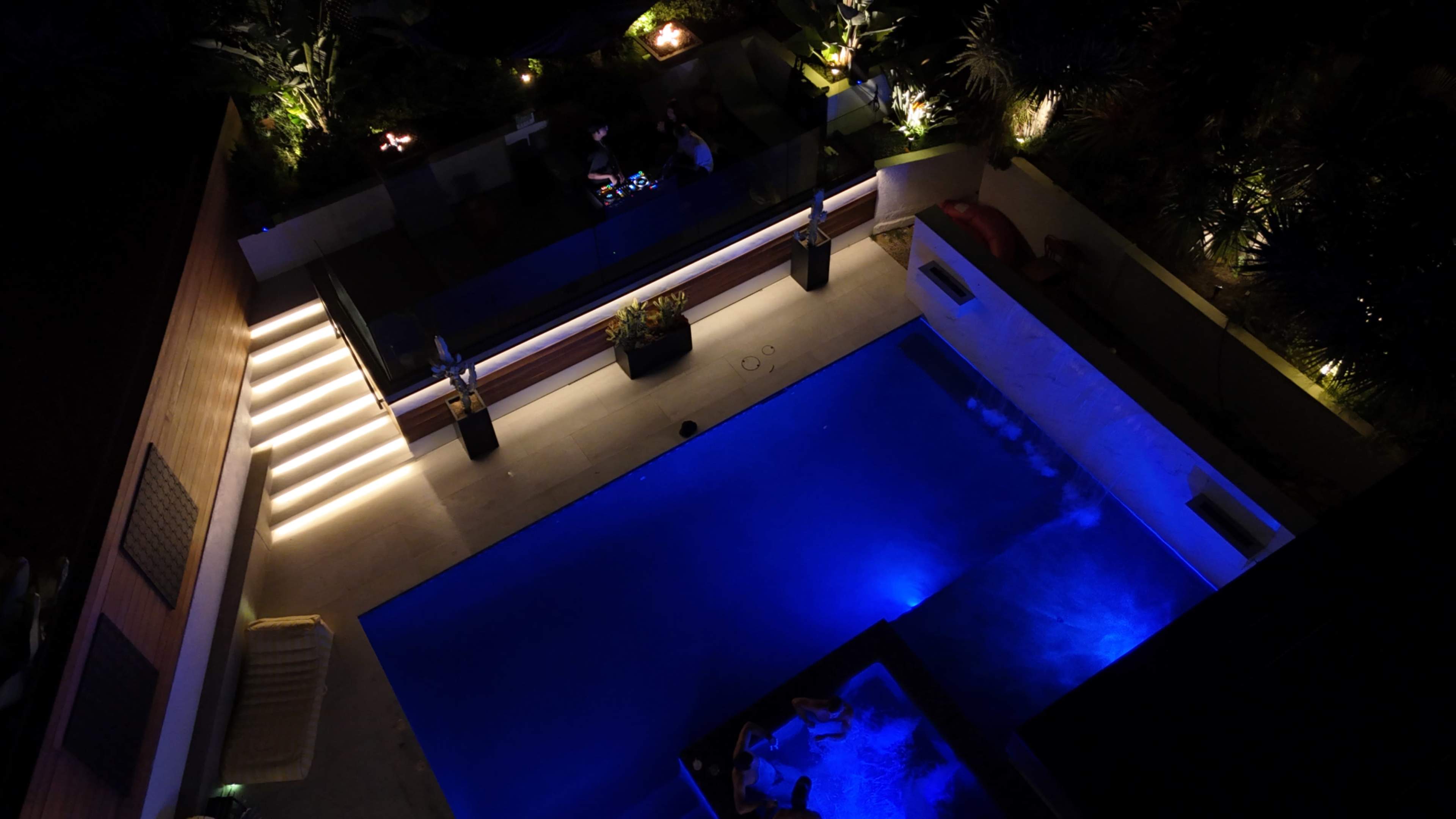 The image shows a well-lit outdoor pool area at night, featuring a blue swimming pool, surrounding steps, and several plants and seating arrangements.