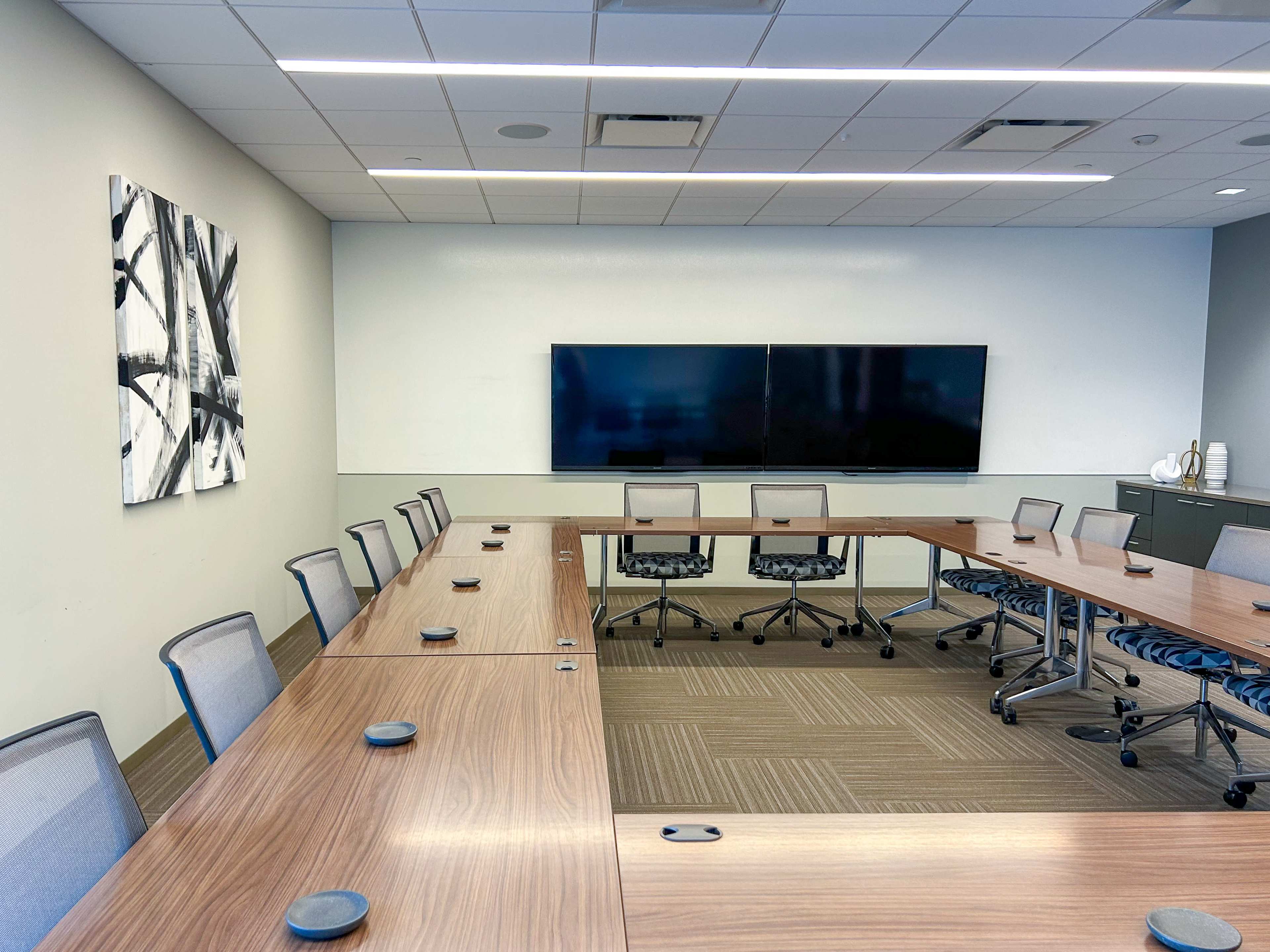 The image shows a well-lit conference room with a long table surrounded by chairs, equipped with two large screens on the wall and a modern artwork on the side.