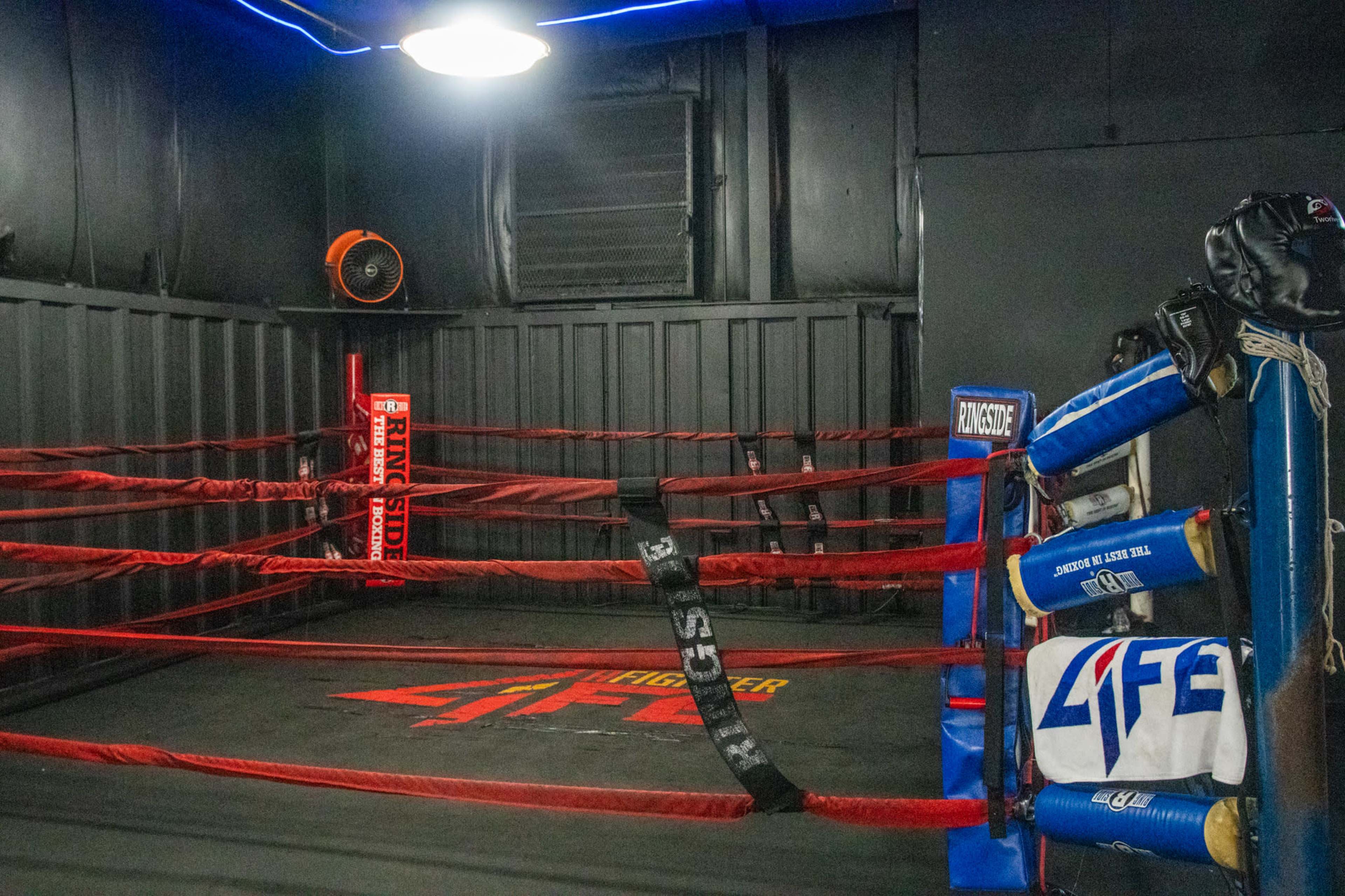 The image shows a boxing ring in a dimly lit gym, featuring red ropes, a fan, and various boxing equipment arranged nearby.