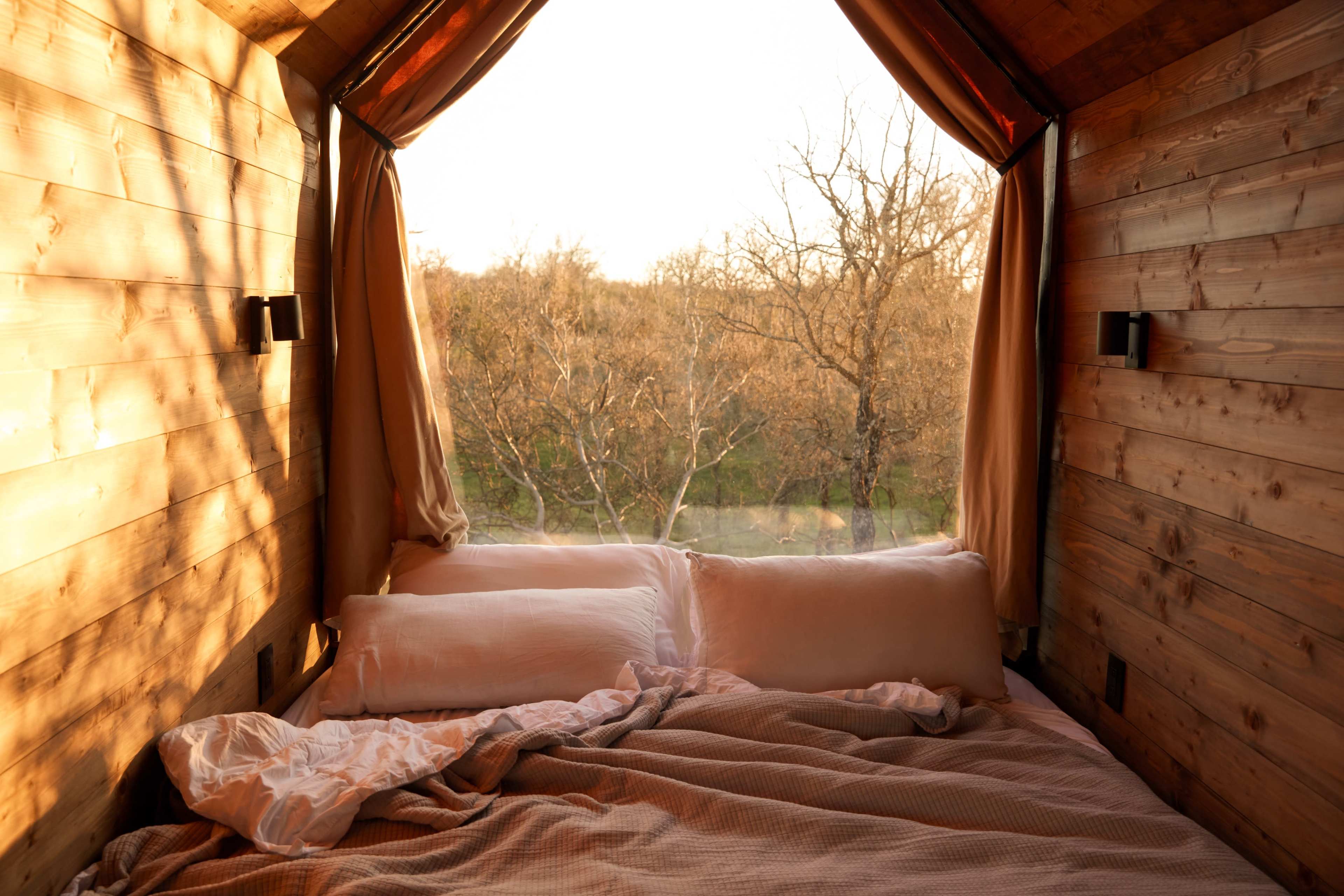 A cozy wooden interior of a cabin features a bed with pillows and a large window opening to a view of trees outside.