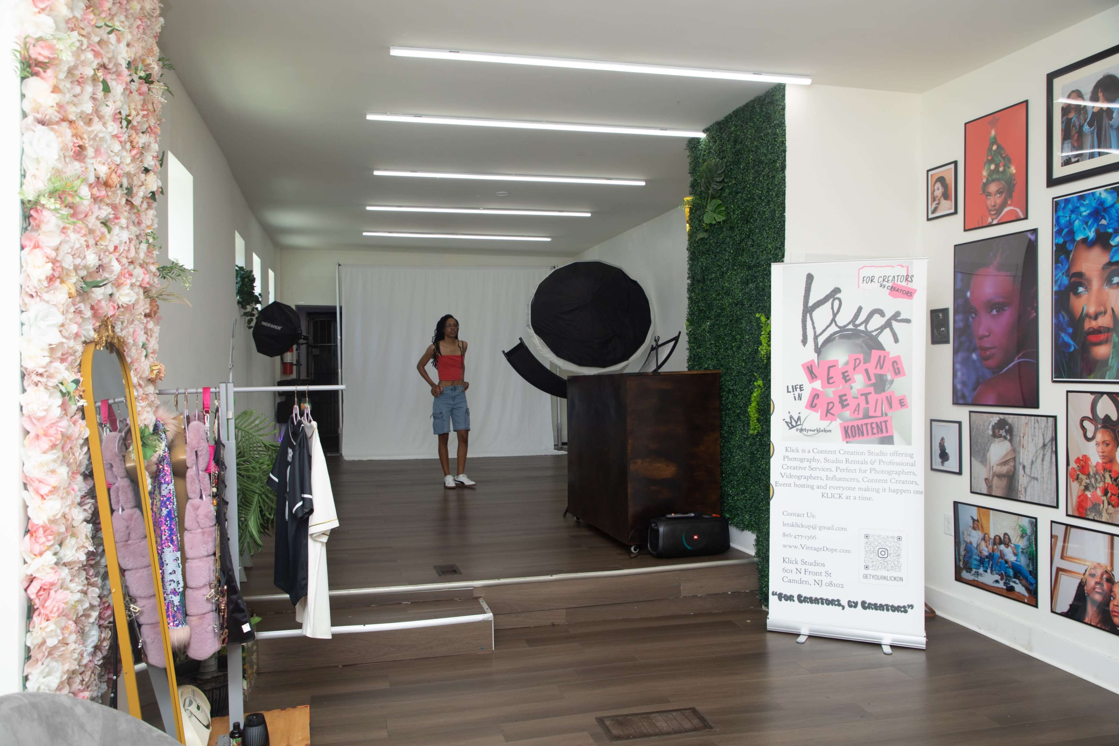The image shows a well-lit studio space featuring a model standing in front of a backdrop, surrounded by decorative walls and framed photographs.