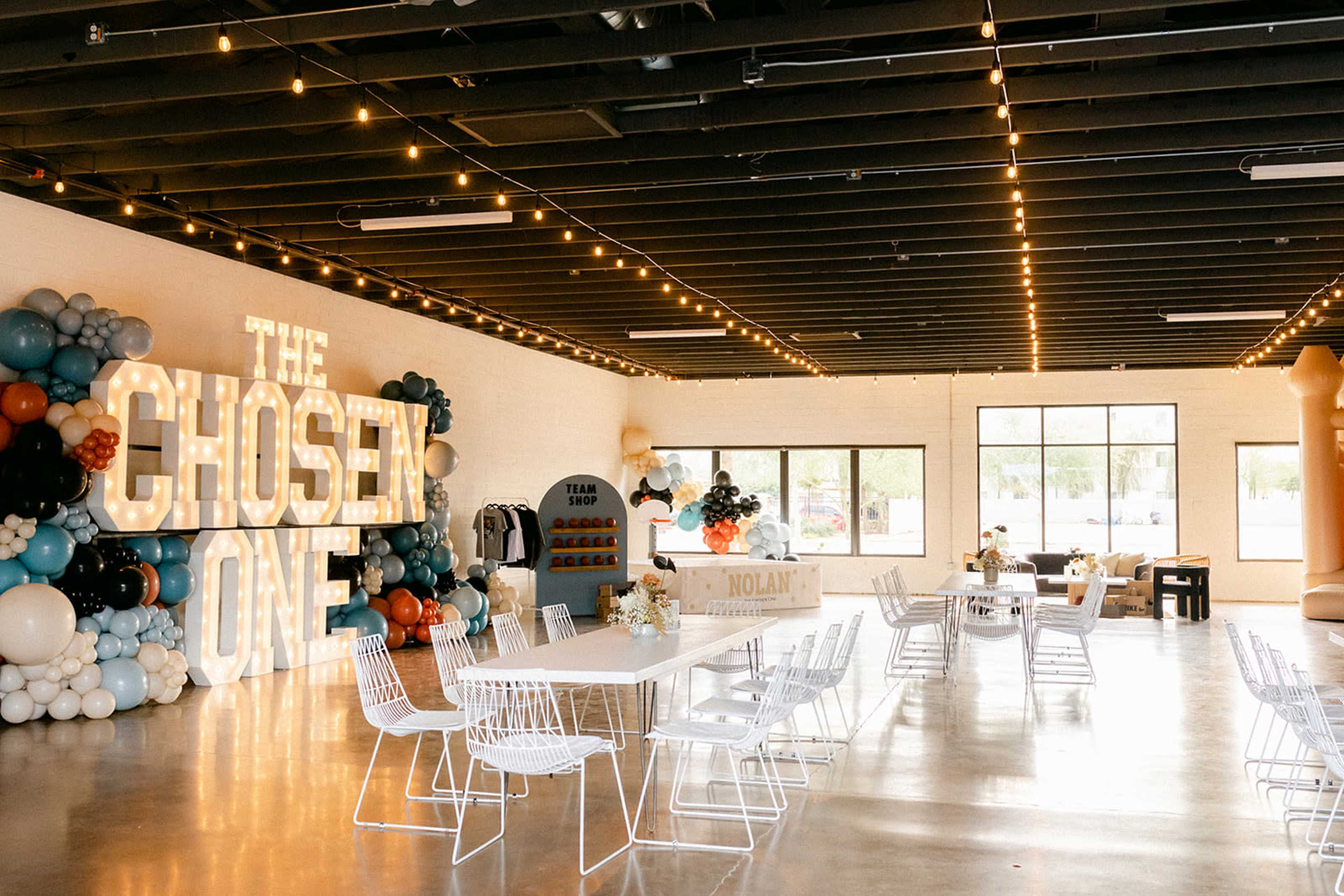 The interior of a spacious event venue features large illuminated letters spelling "THE CHOSEN ONE," surrounded by decorative balloons and white metal chairs arranged around tables.