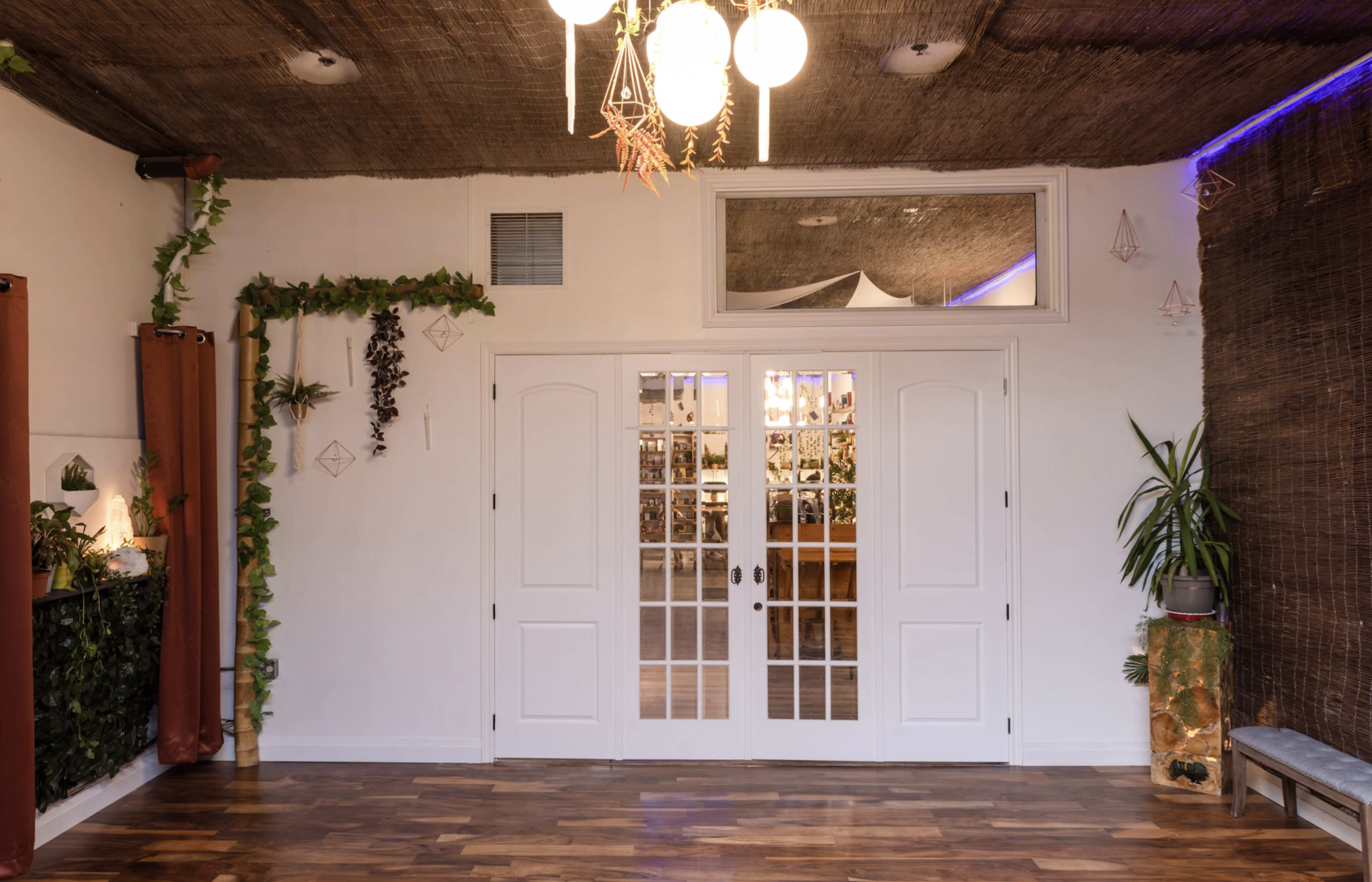 The image shows a bright indoor space featuring double doors, a wooden floor, and decor elements like plants and a woven ceiling.