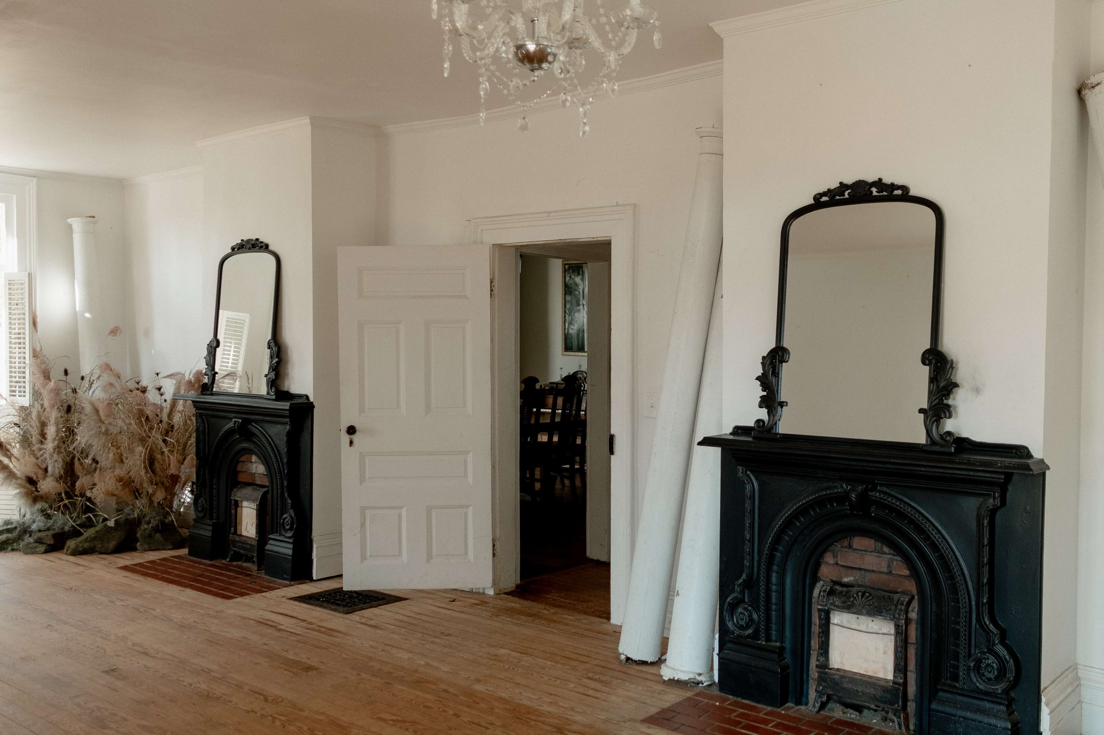The room features two black ornate fireplaces with mirrors above them, an open door leading to another space, and wooden flooring.