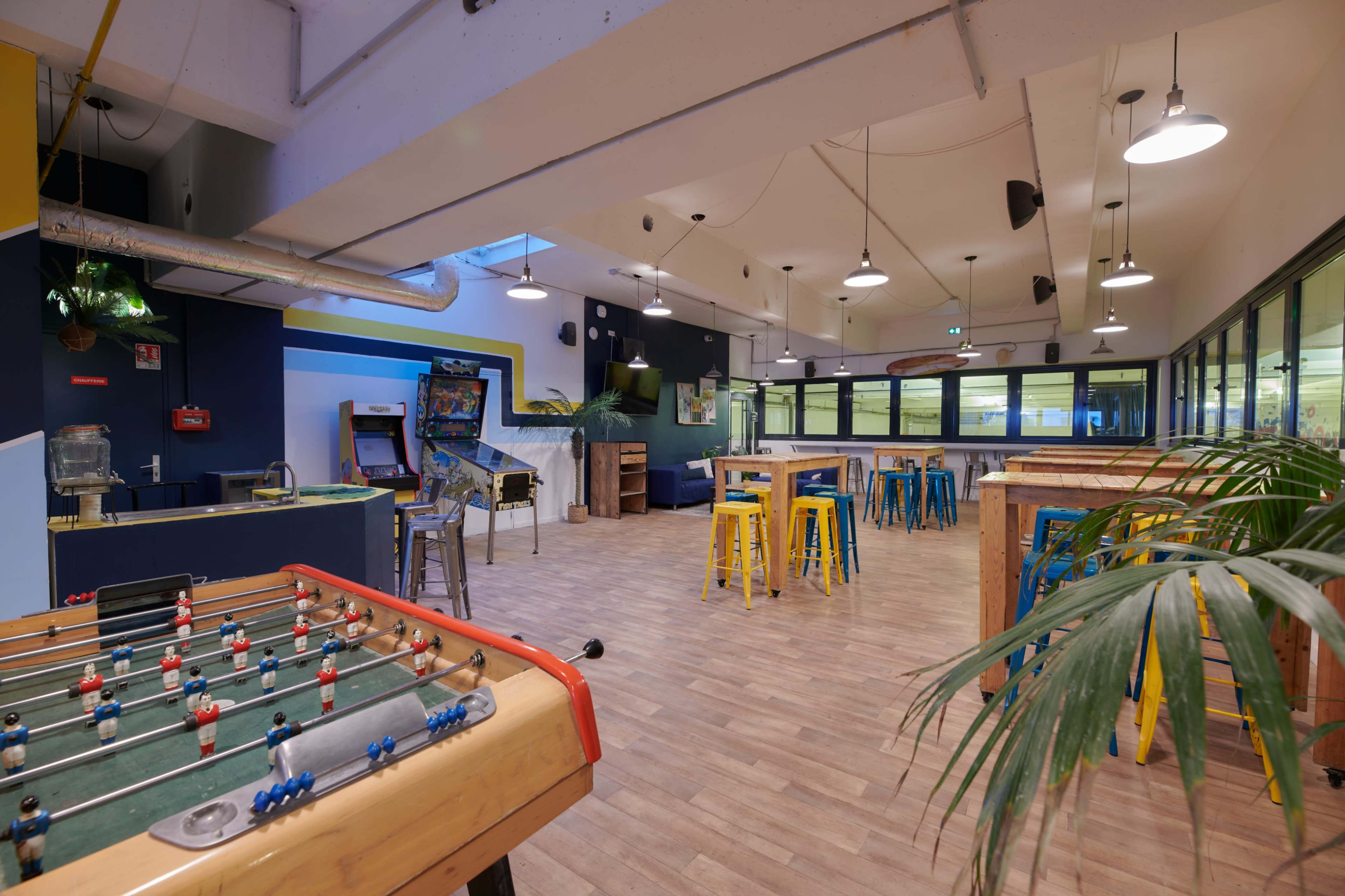 A spacious recreational area features a foosball table, colorful bar stools, wooden tables, and arcade machines.