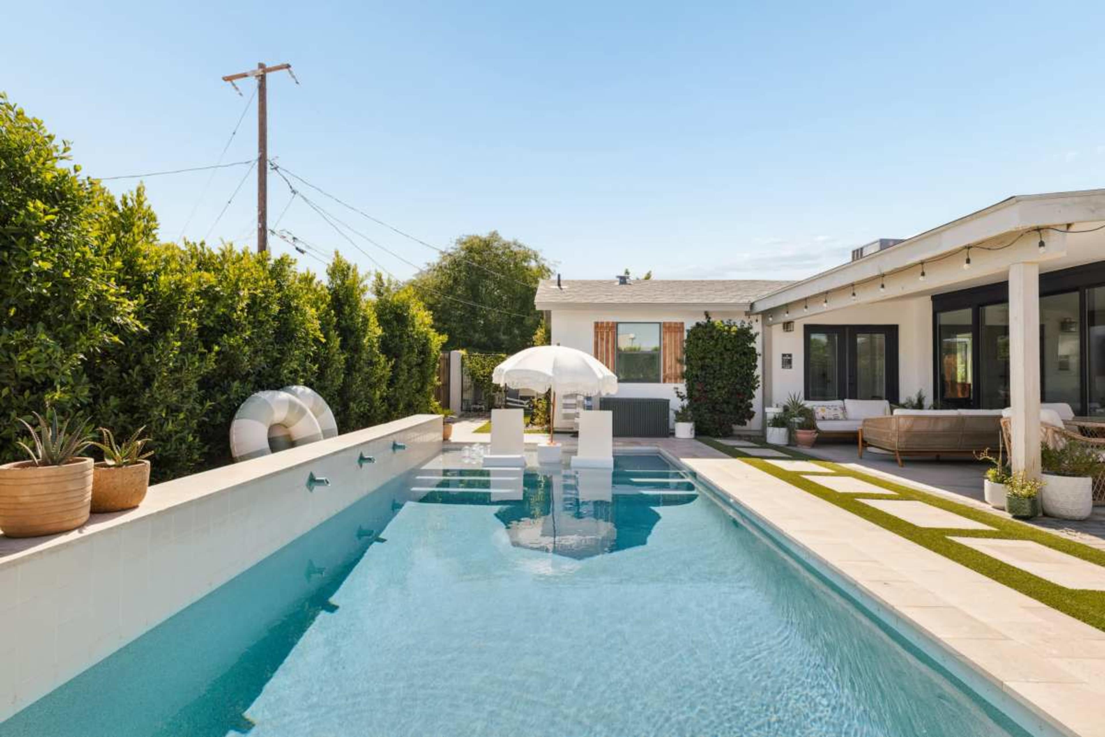 The image shows a modern backyard featuring a swimming pool with a white umbrella, surrounded by neatly trimmed greenery and a stylish house with large windows.