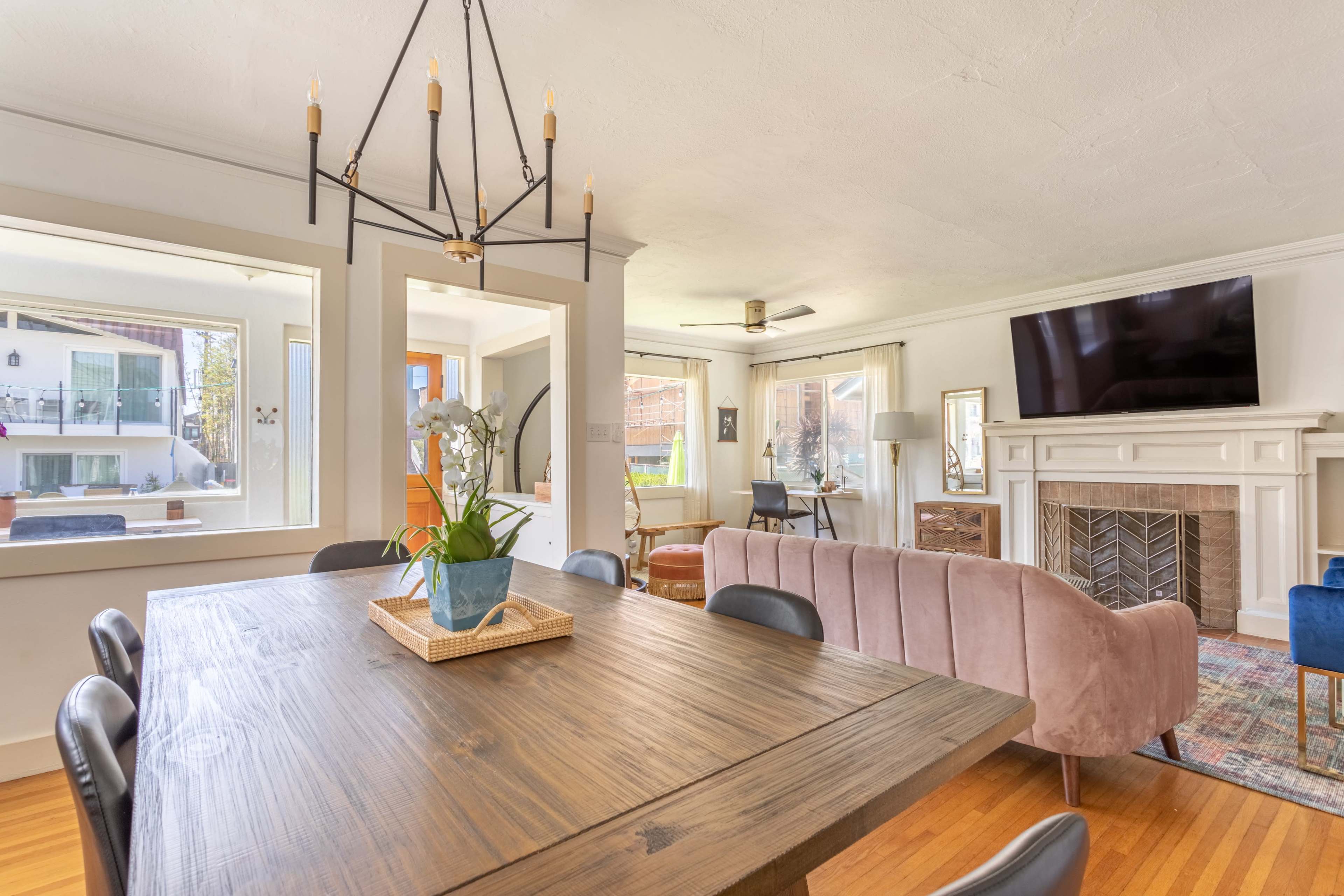 A spacious living and dining area featuring a wooden dining table, a pink sofa, a television mounted on the wall, and a decorative fireplace.